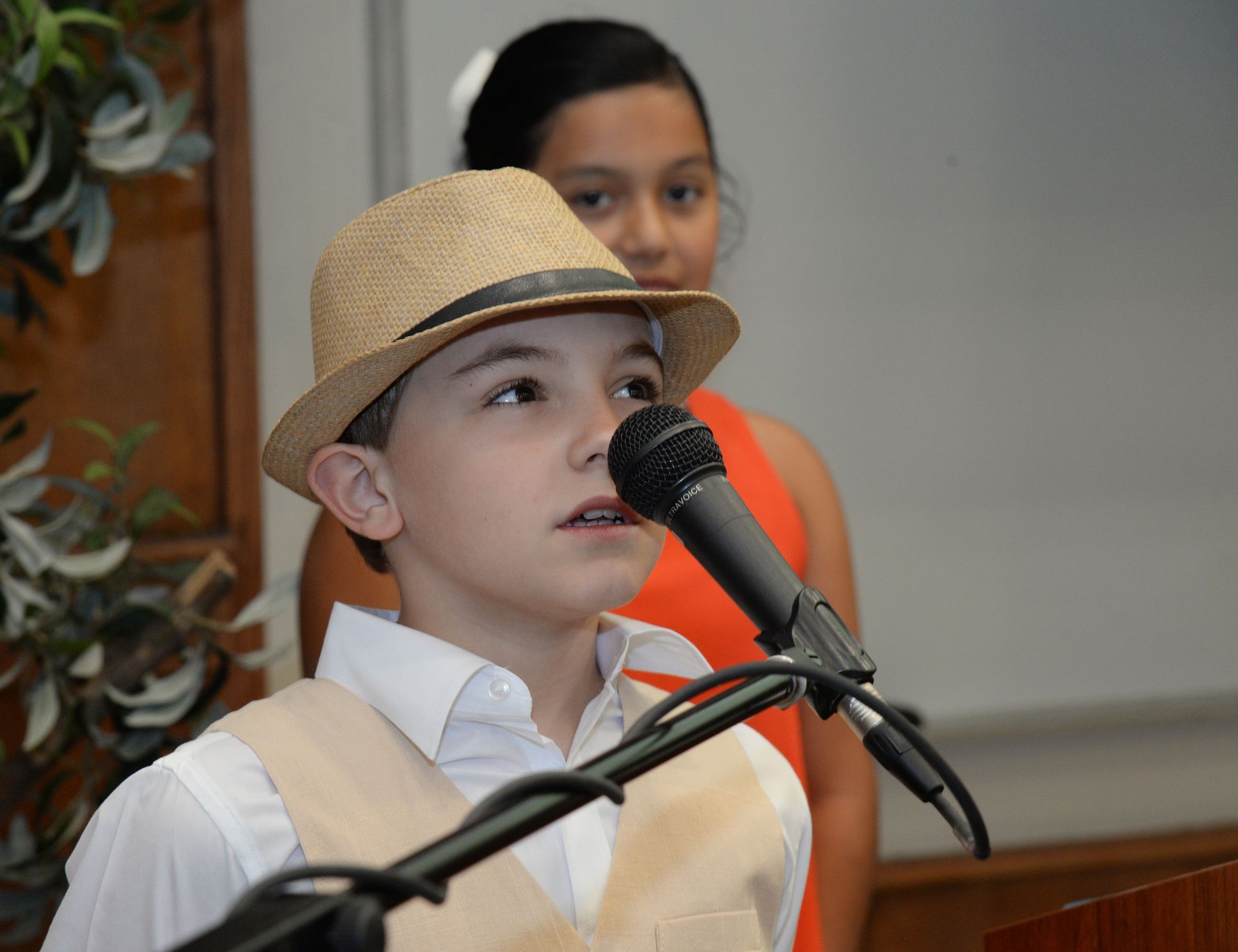 Boy in hat speaks into microphone; another child in background.