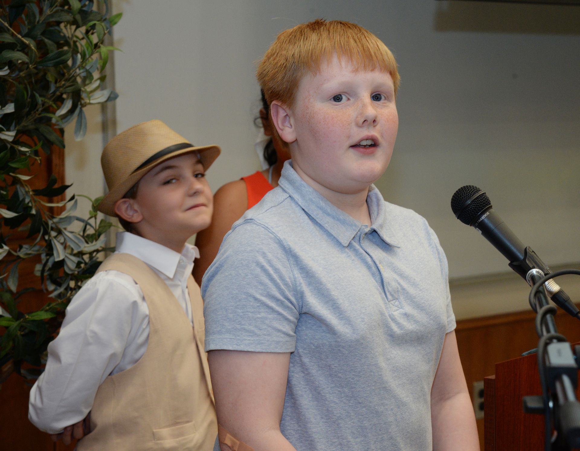 Boy speaking into a microphone. Another boy in a hat stands behind him. Indoors, presumably a presentation.