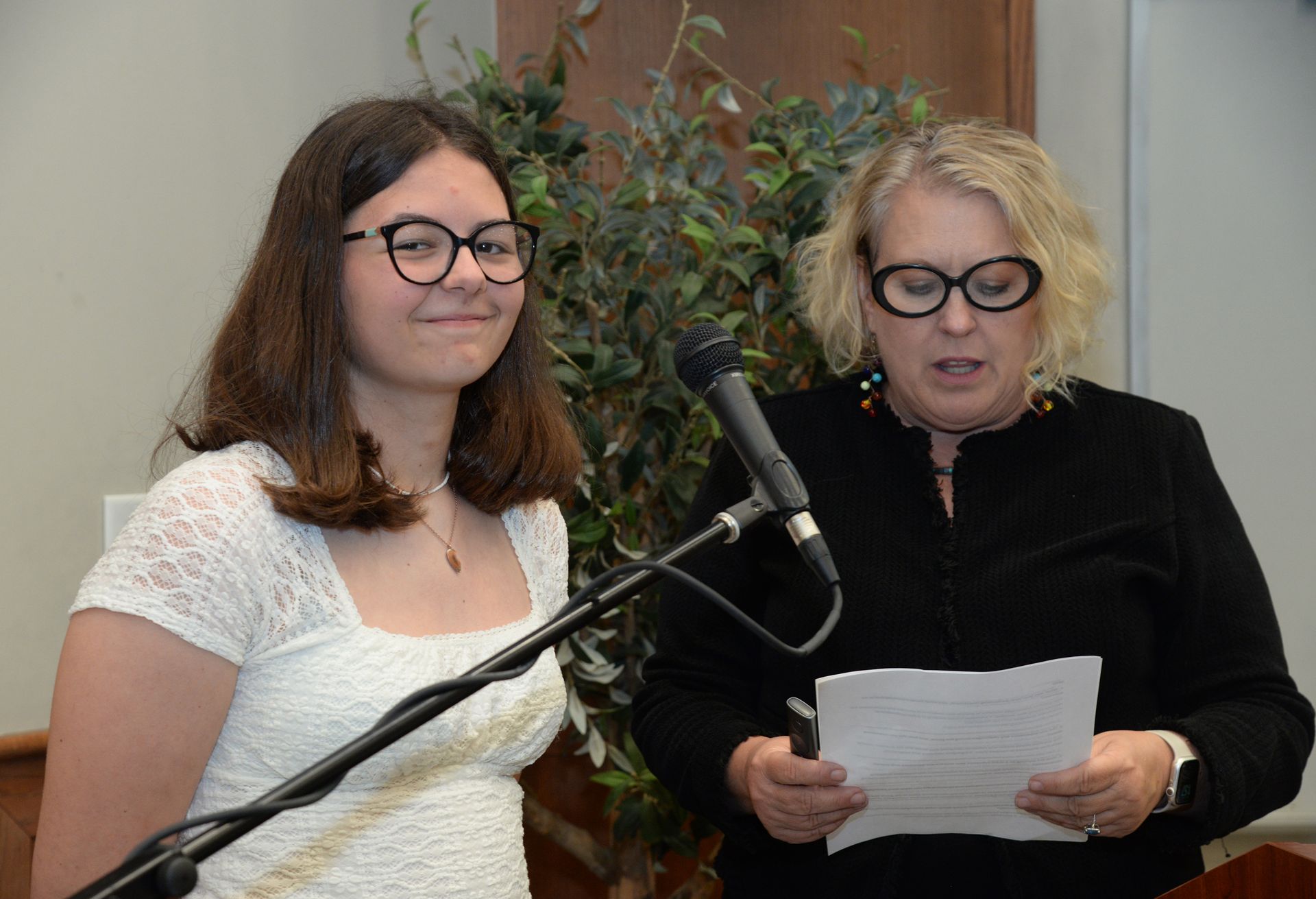 Woman speaking at a microphone, holding papers, and another woman standing beside her, smiling, in front of a plant.