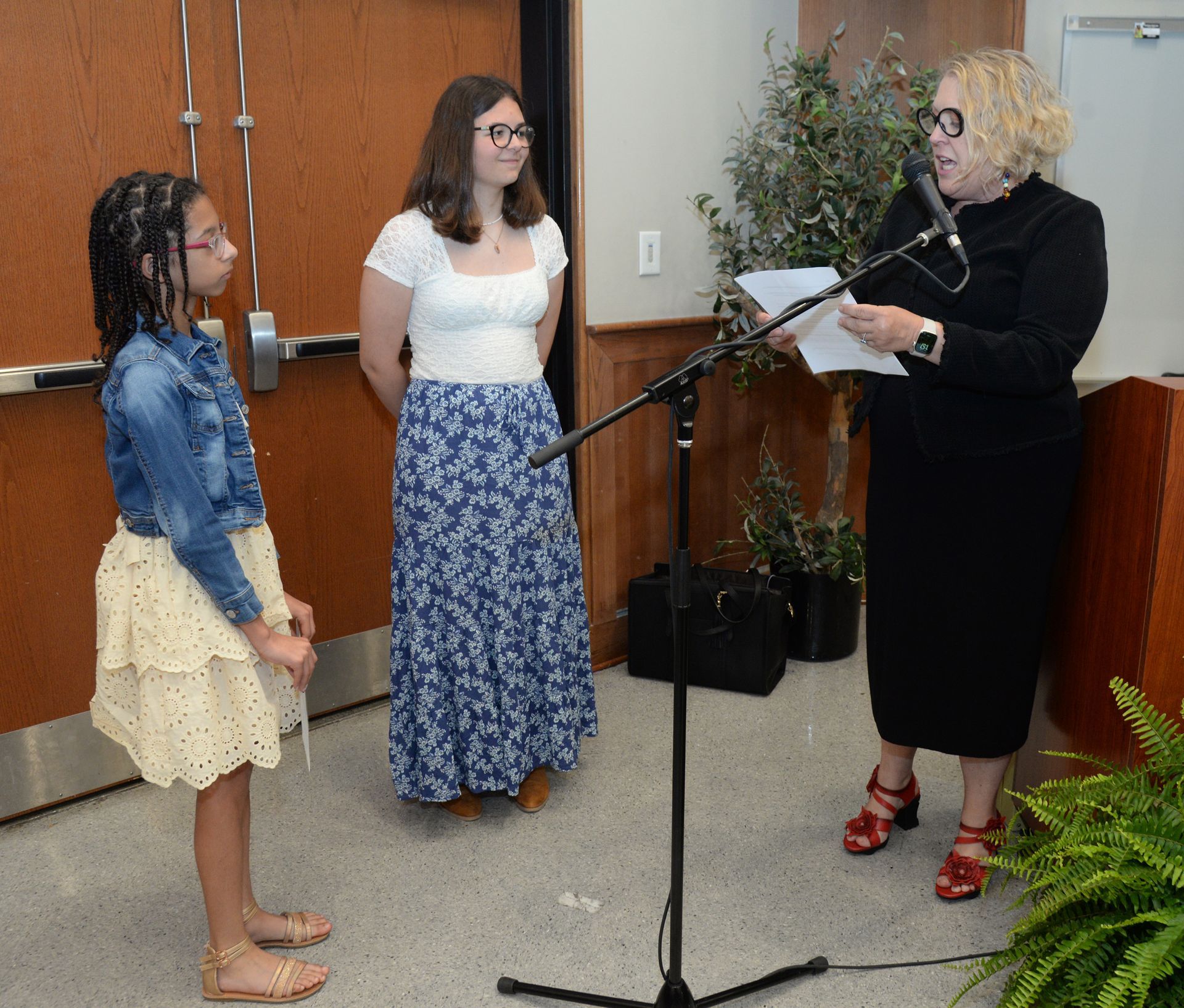 Woman speaking into a microphone, two others stand nearby. Building interior.