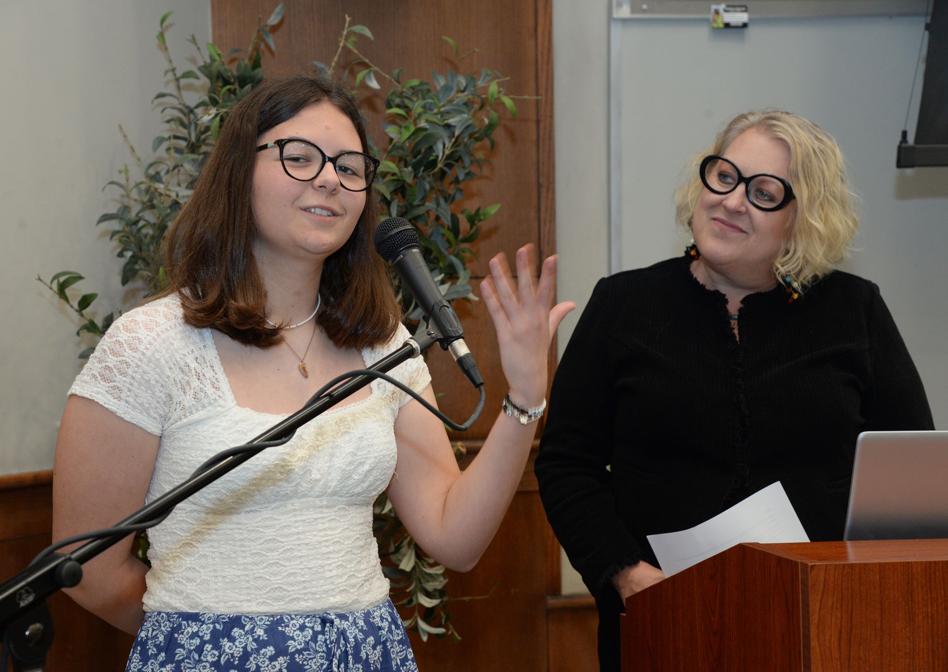 Woman speaking at a lectern, gesturing with hand, another woman stands beside, holding papers. Indoors.
