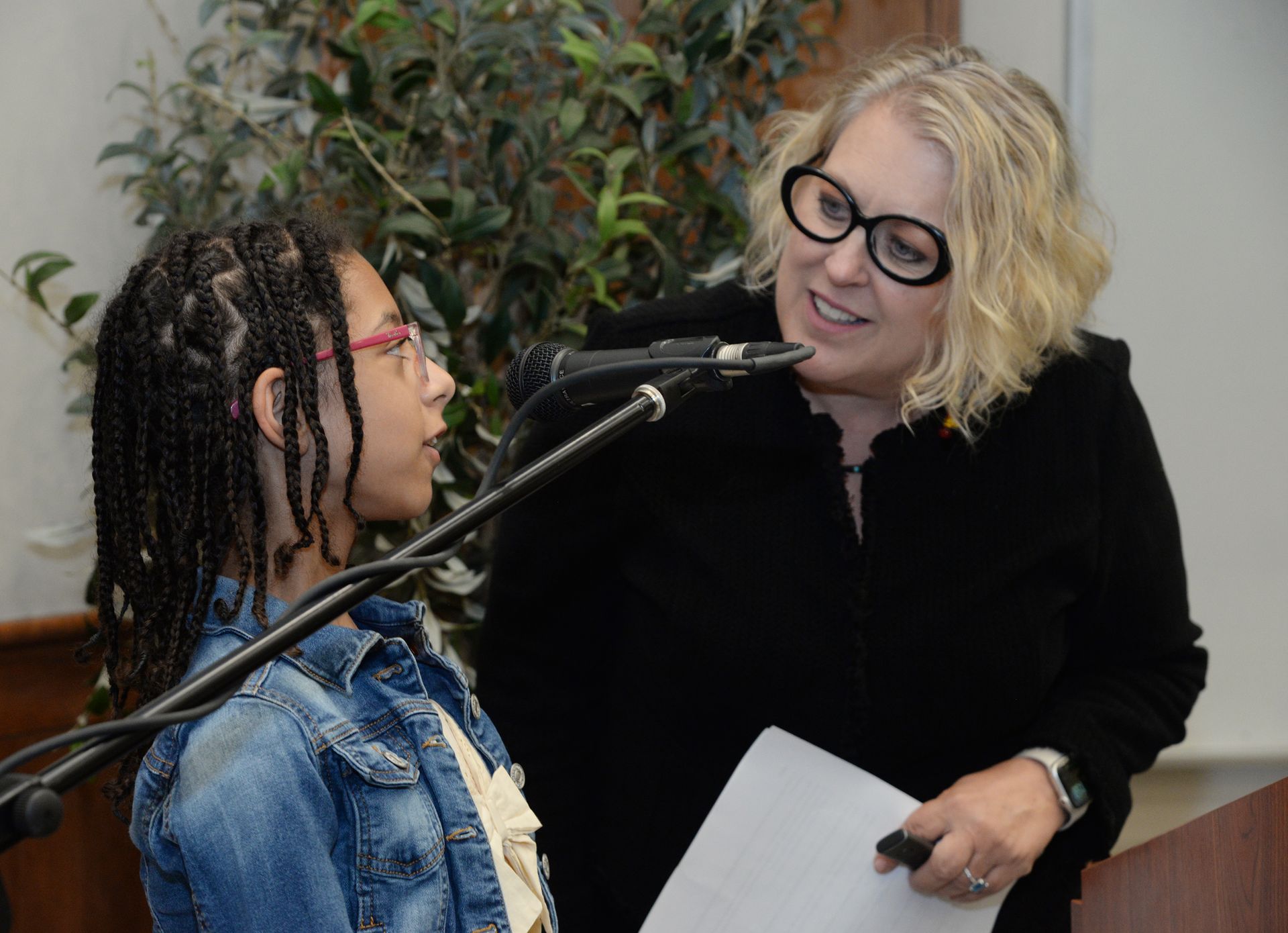 Woman with glasses leans toward a girl with braids, speaking into a microphone. Both are in front of a podium.