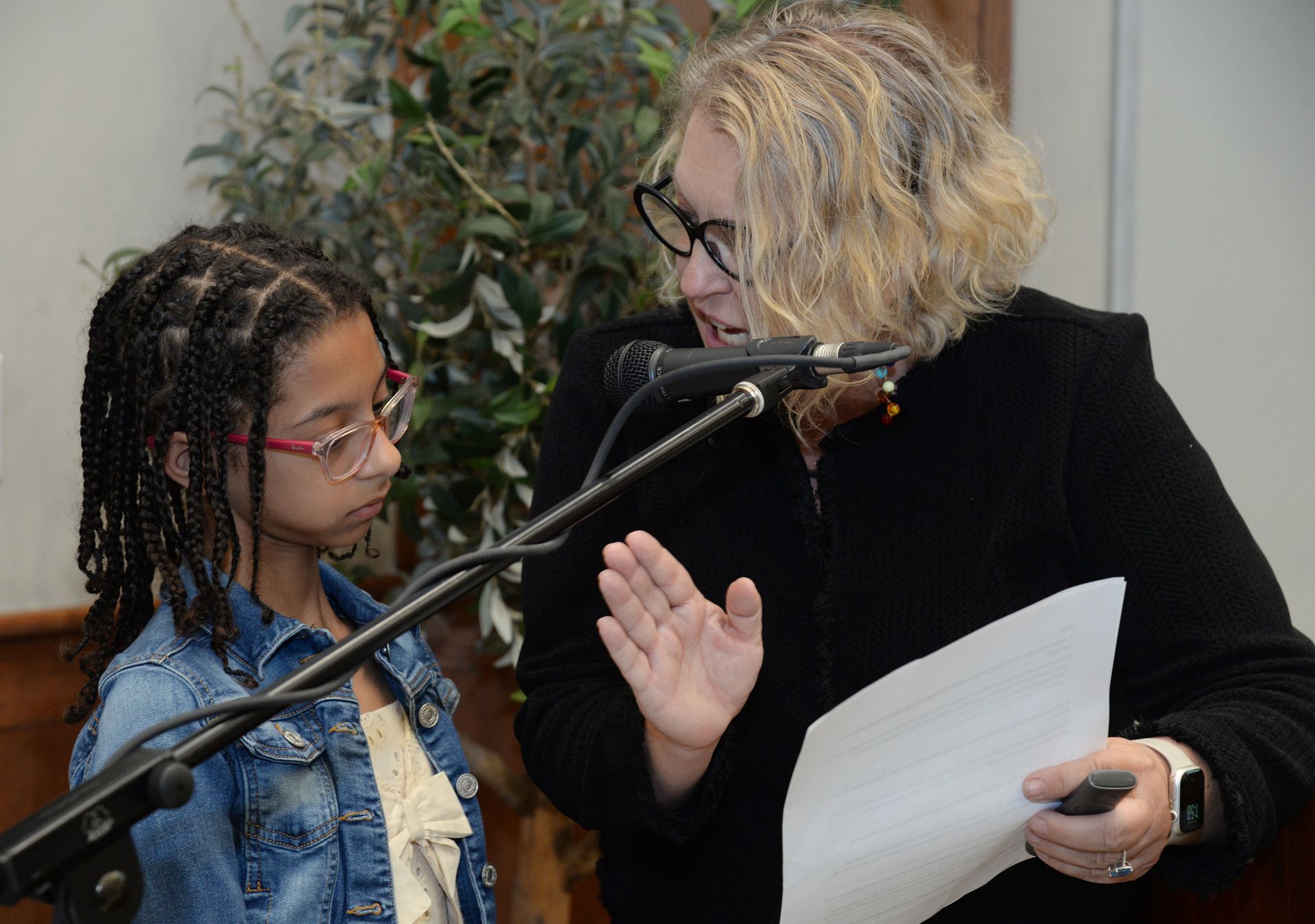 A woman with a microphone gestures toward a child; both stand at a podium.
