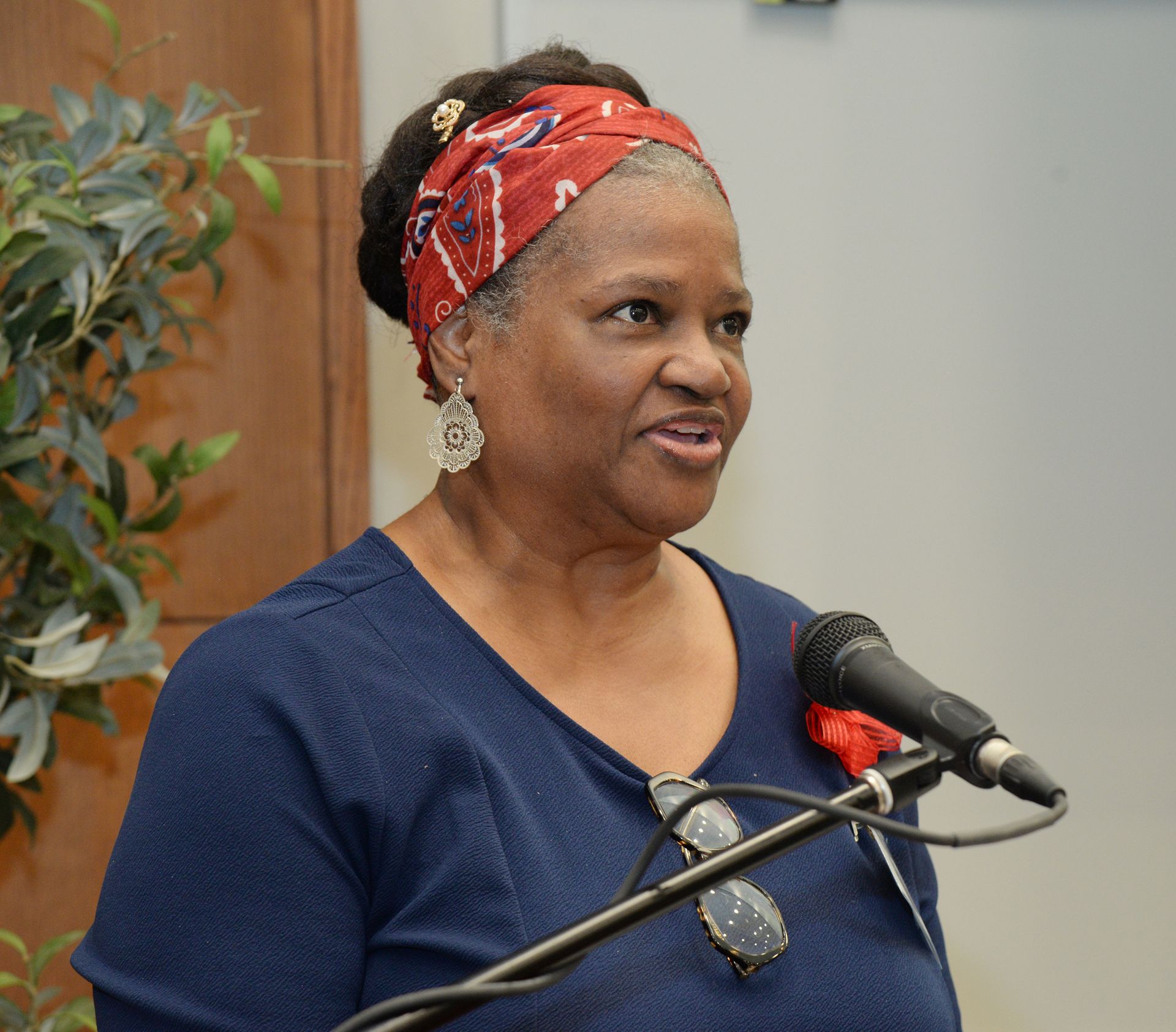 Woman speaking at a microphone, wearing a red headscarf and blue top. She is indoors with a plant in the background.
