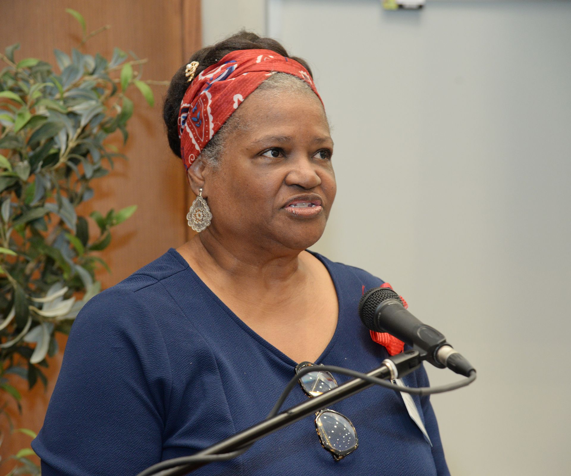 Woman speaking at a microphone, wearing a blue shirt and patterned headscarf.