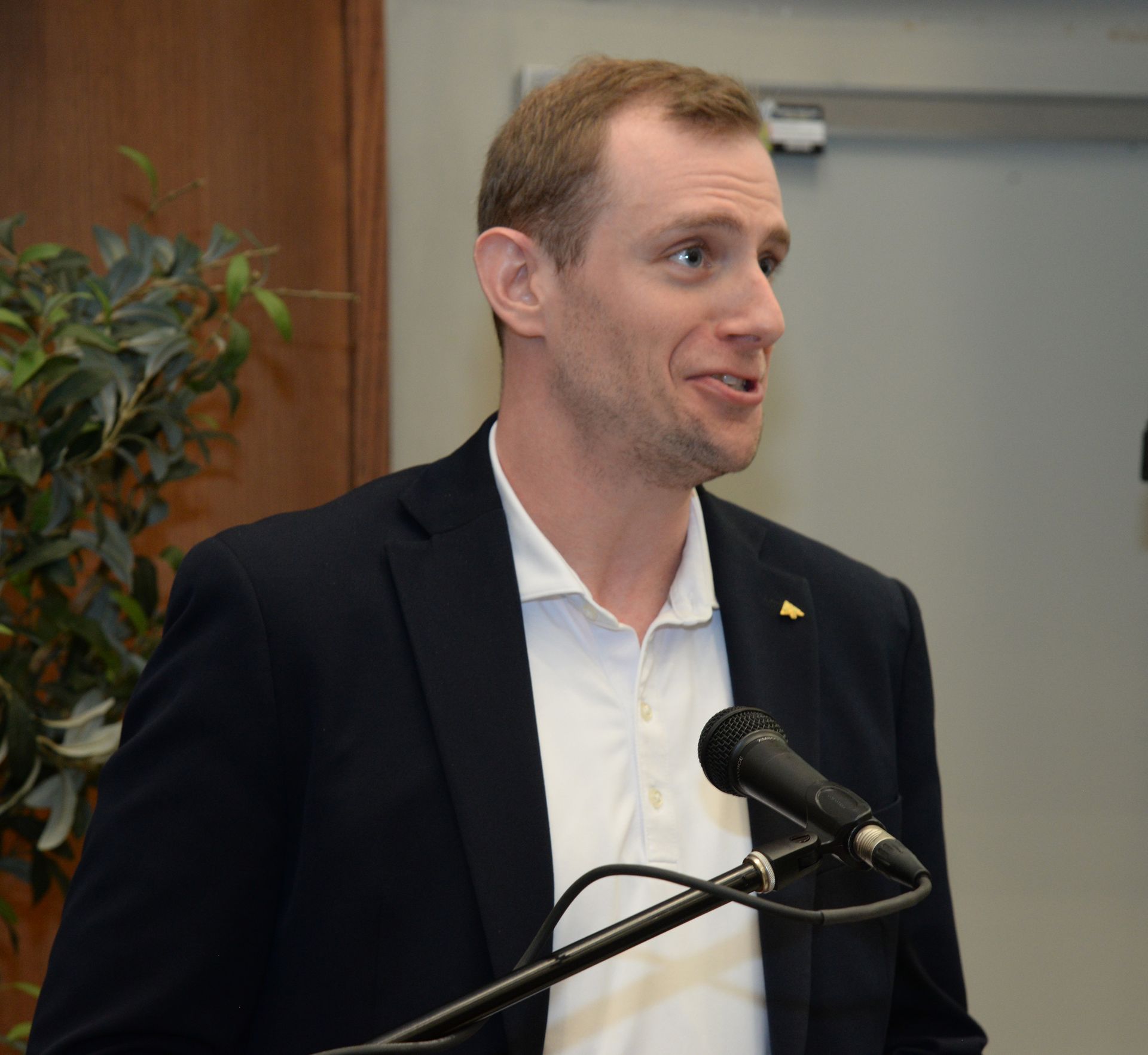 Man in a navy blazer and white polo shirt speaking into a microphone.