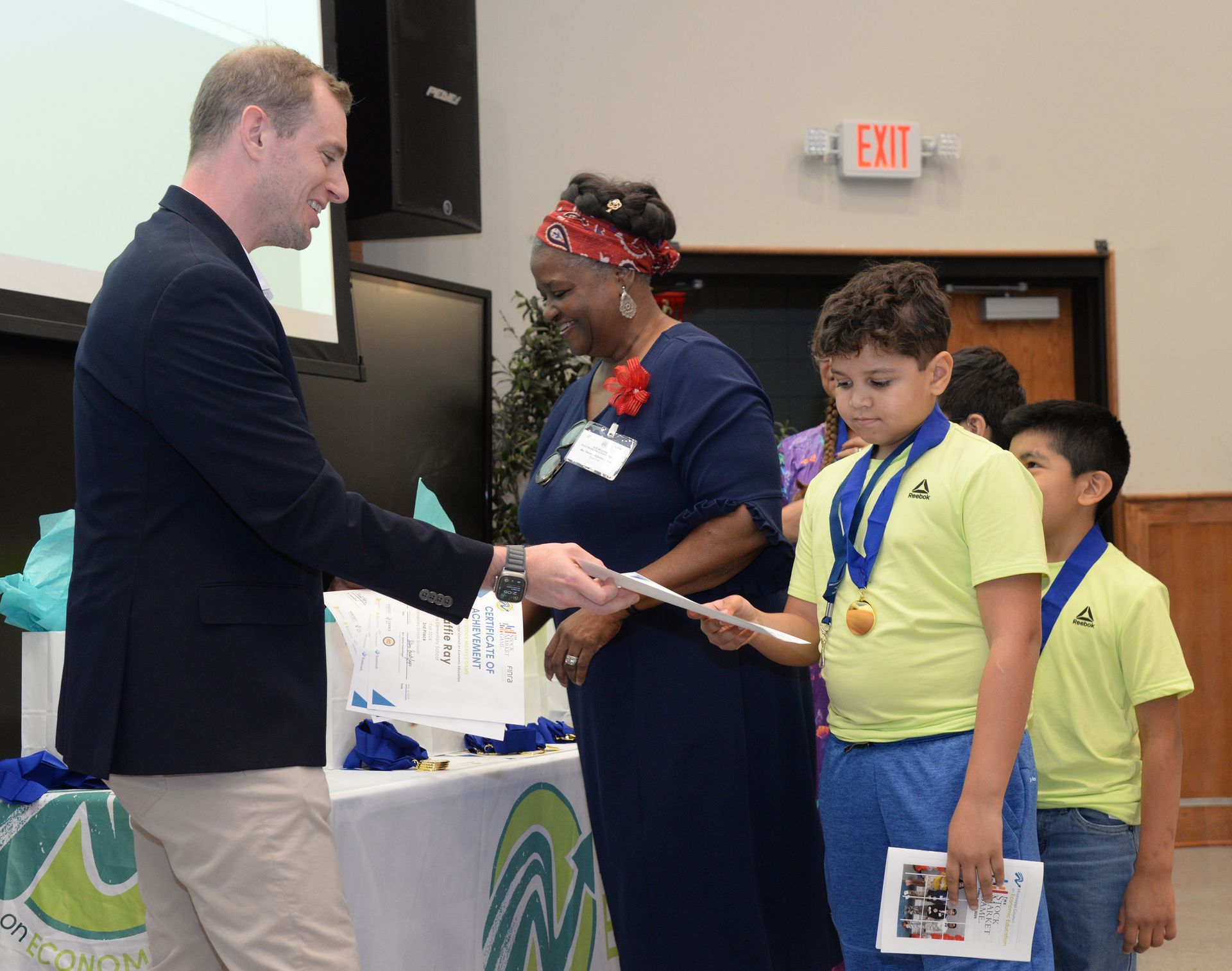 Man presenting certificate to a boy wearing a medal; woman watches. Others stand nearby in an event space.