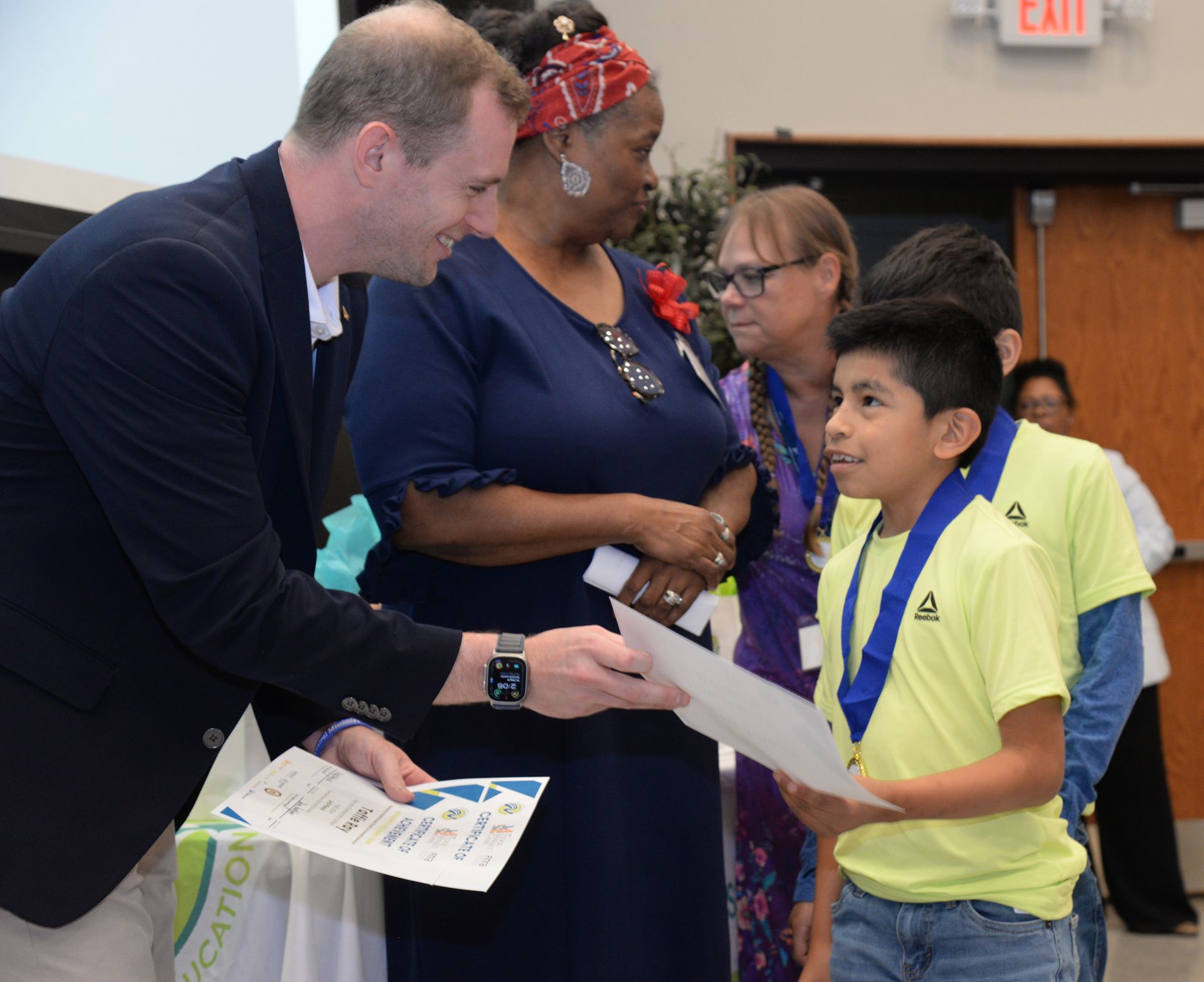 Man handing certificate to boy wearing a medal; others observe in an indoor setting.