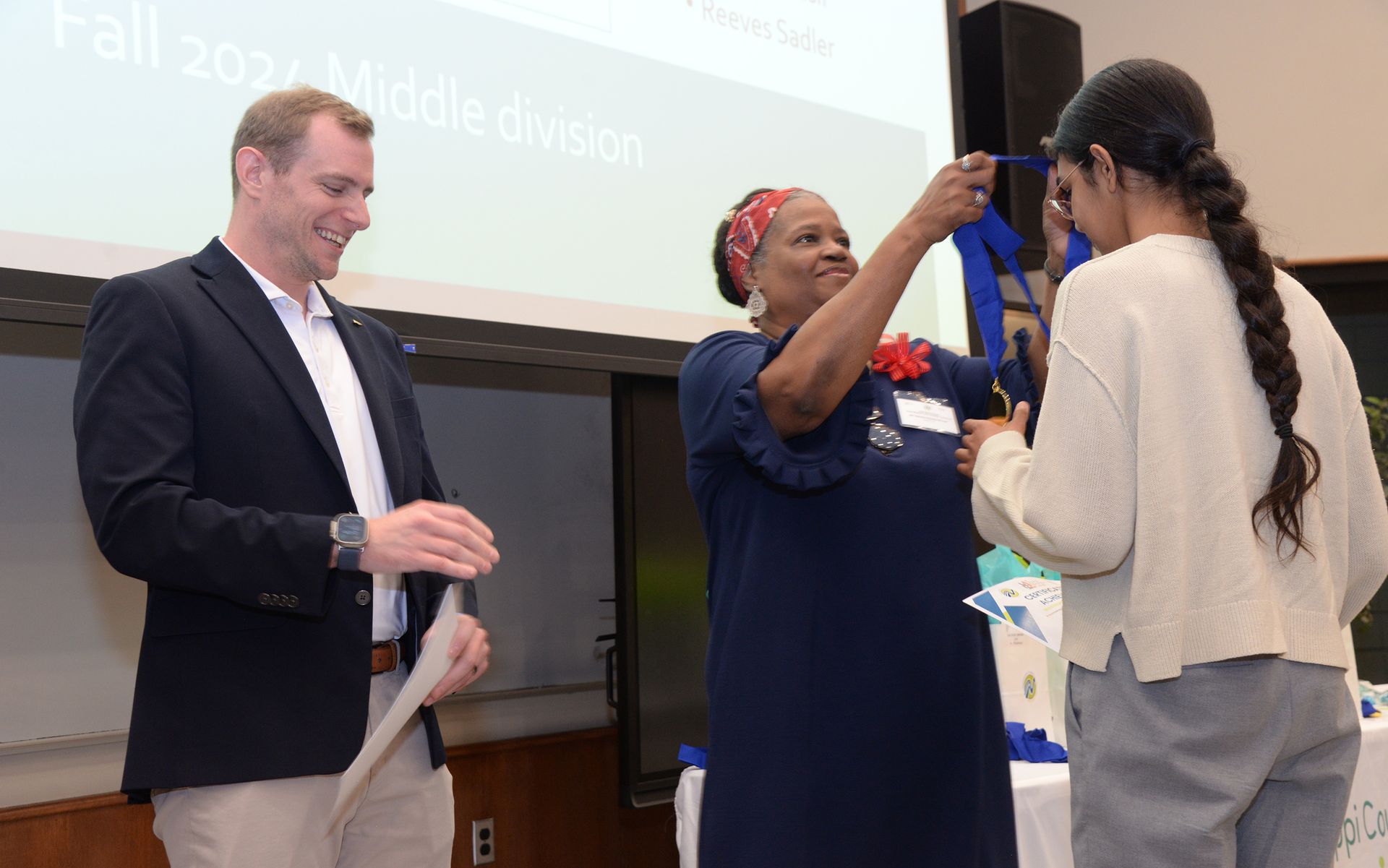 Man and woman presenting a blue sash to another person. Event held in a room with a presentation screen.