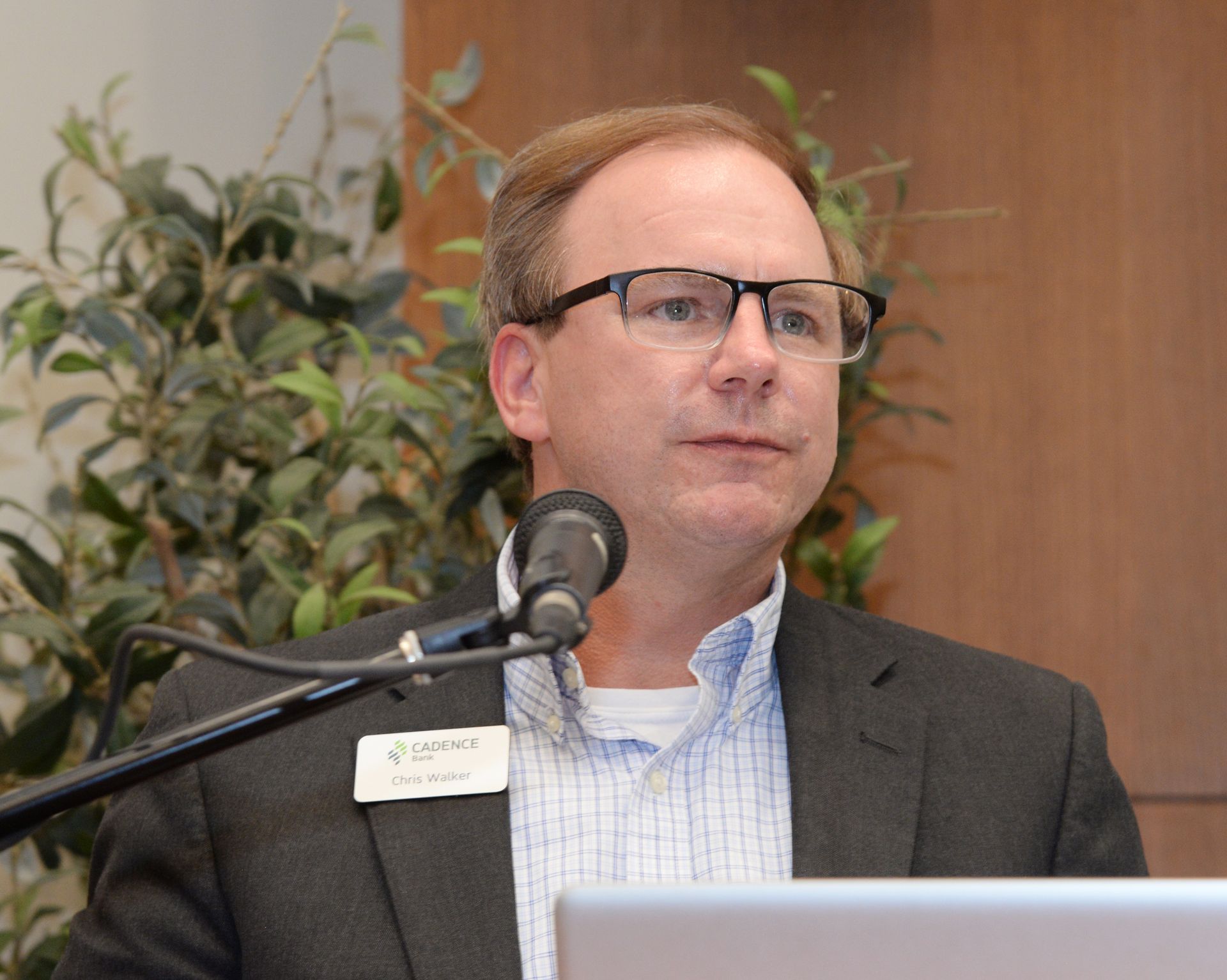 Man with glasses speaks at a podium with a microphone; presentation in front of greenery.