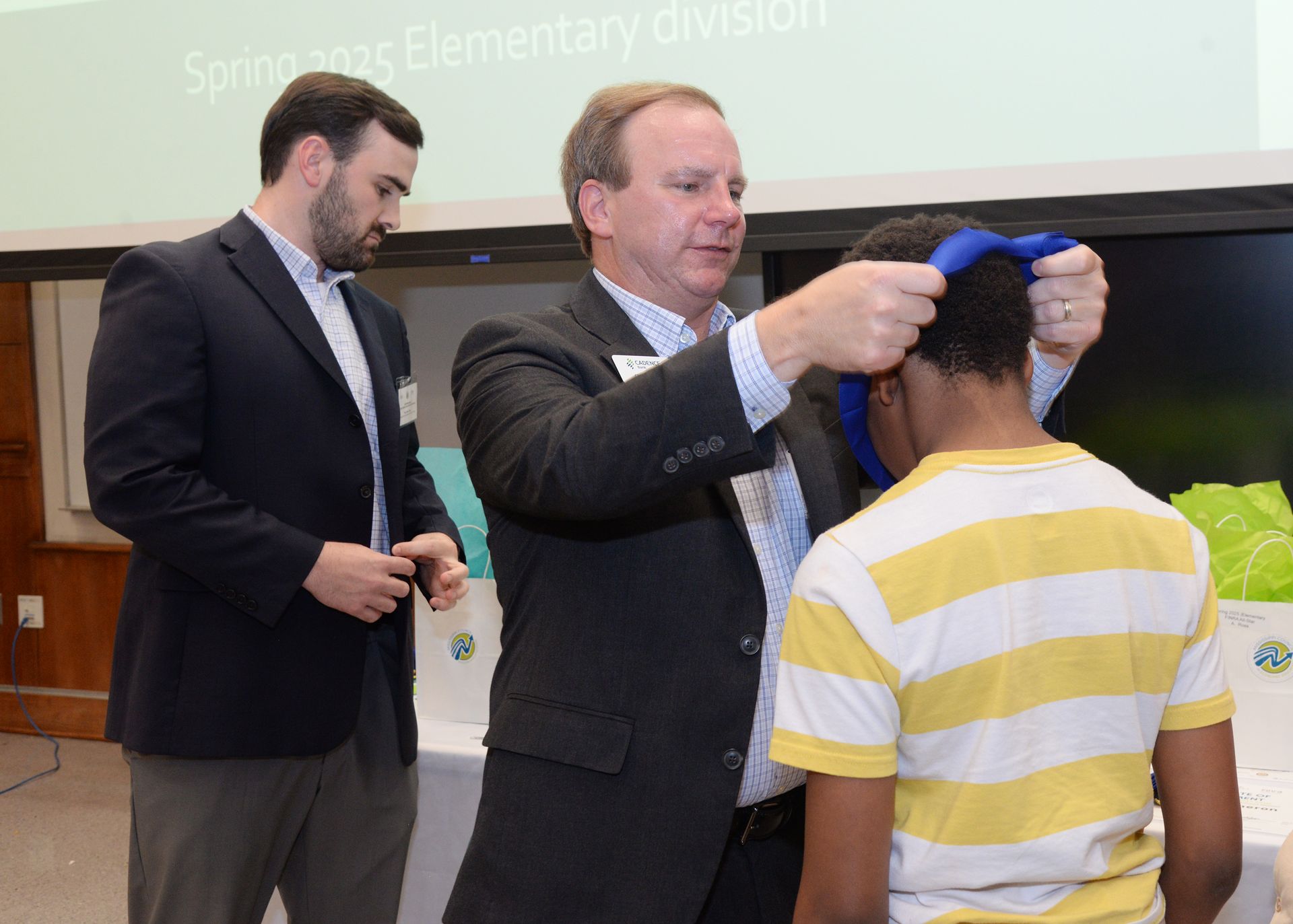 Man places medal on a boy's neck during an awards ceremony. A second man watches.
