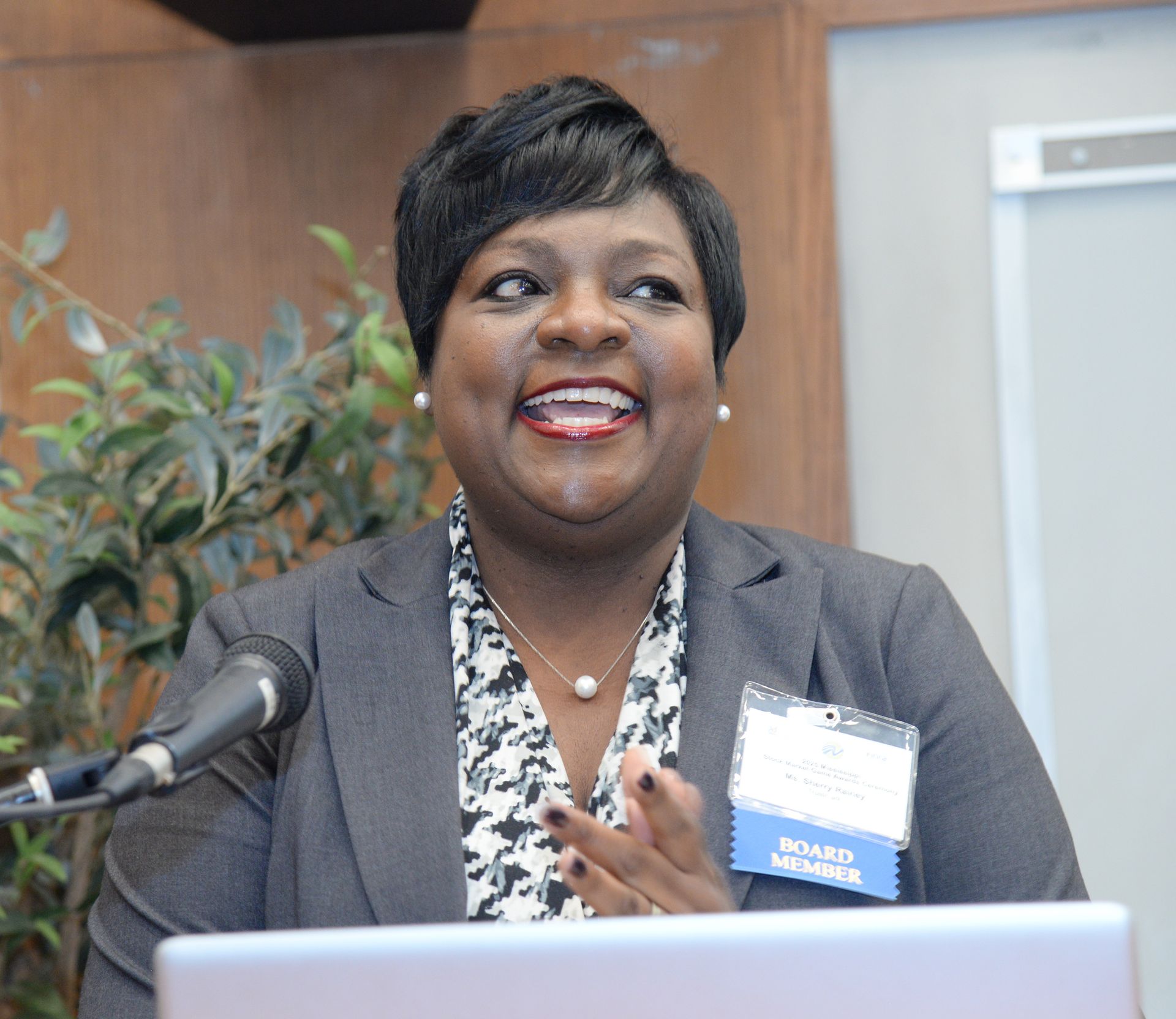 Woman speaking at podium, wearing blazer, smiling, clapping hands.