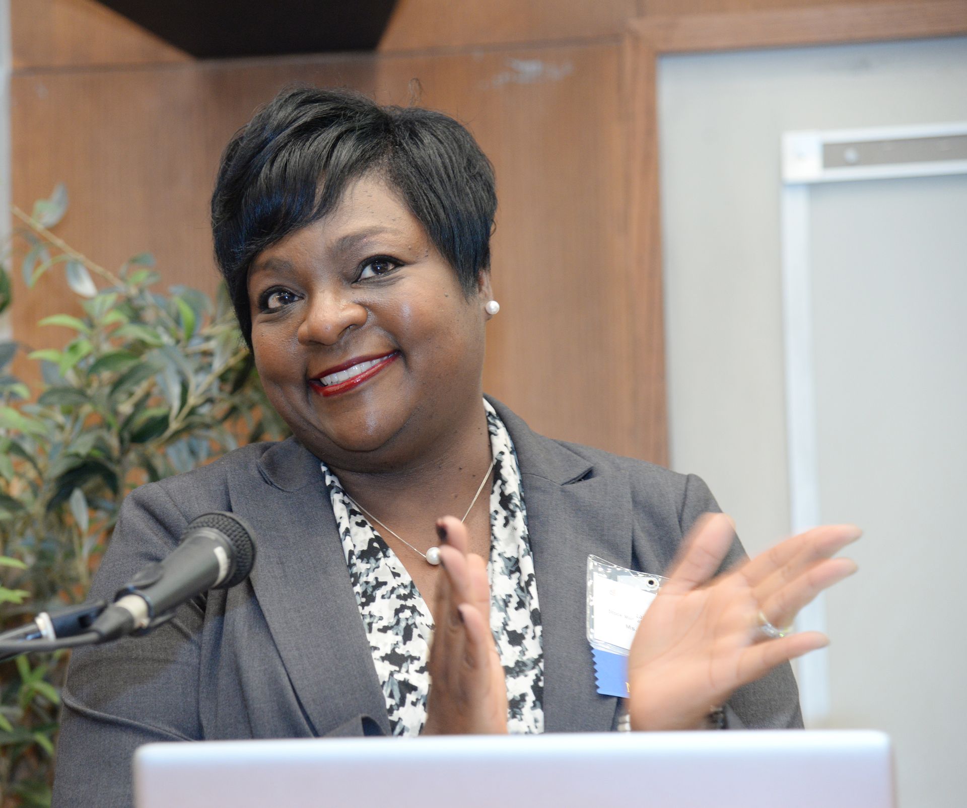Woman in gray suit, applauding at a podium, smiling. A microphone and laptop are visible.