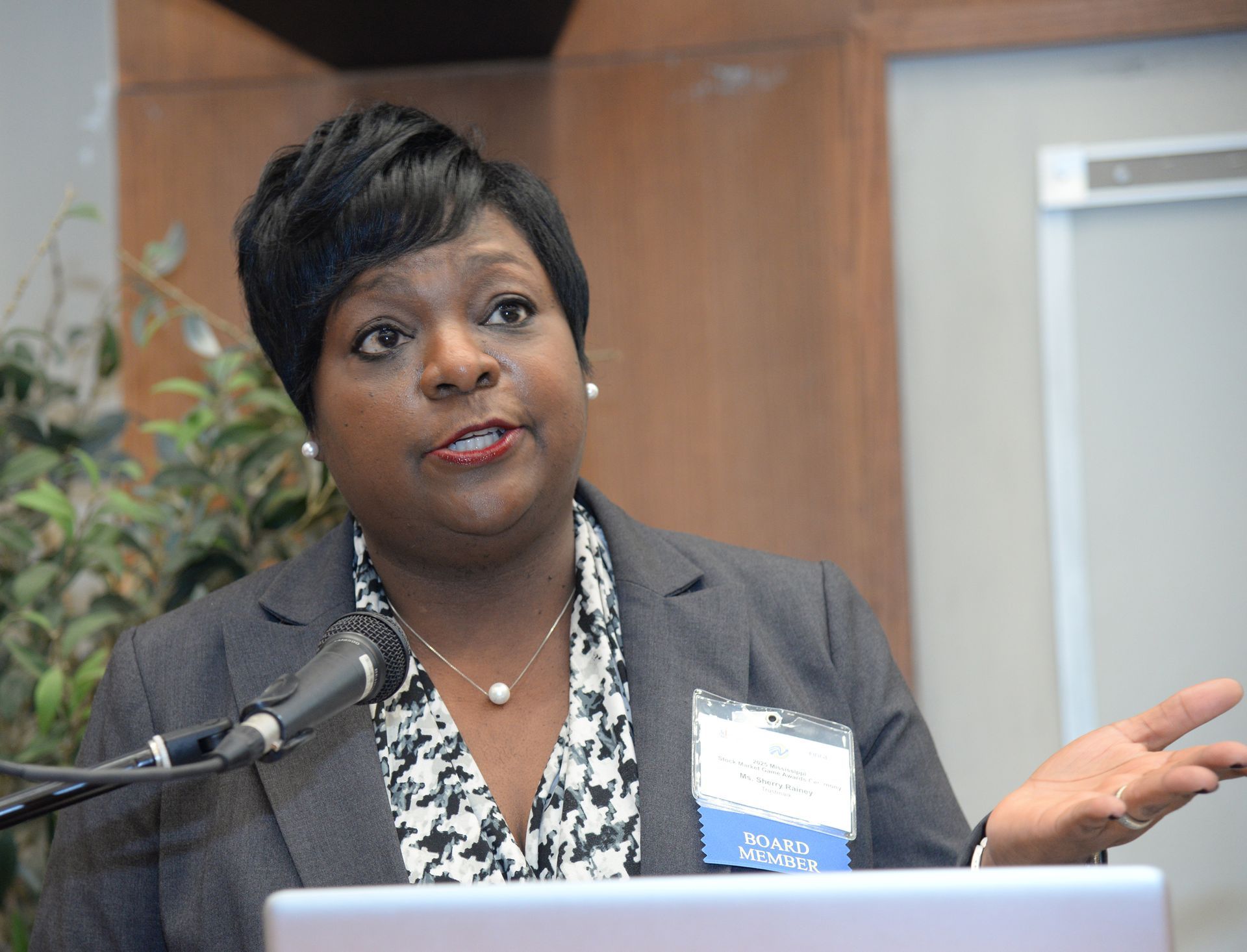 Woman speaking at a conference, gesturing with hand, wearing a blazer over patterned blouse, near a microphone.