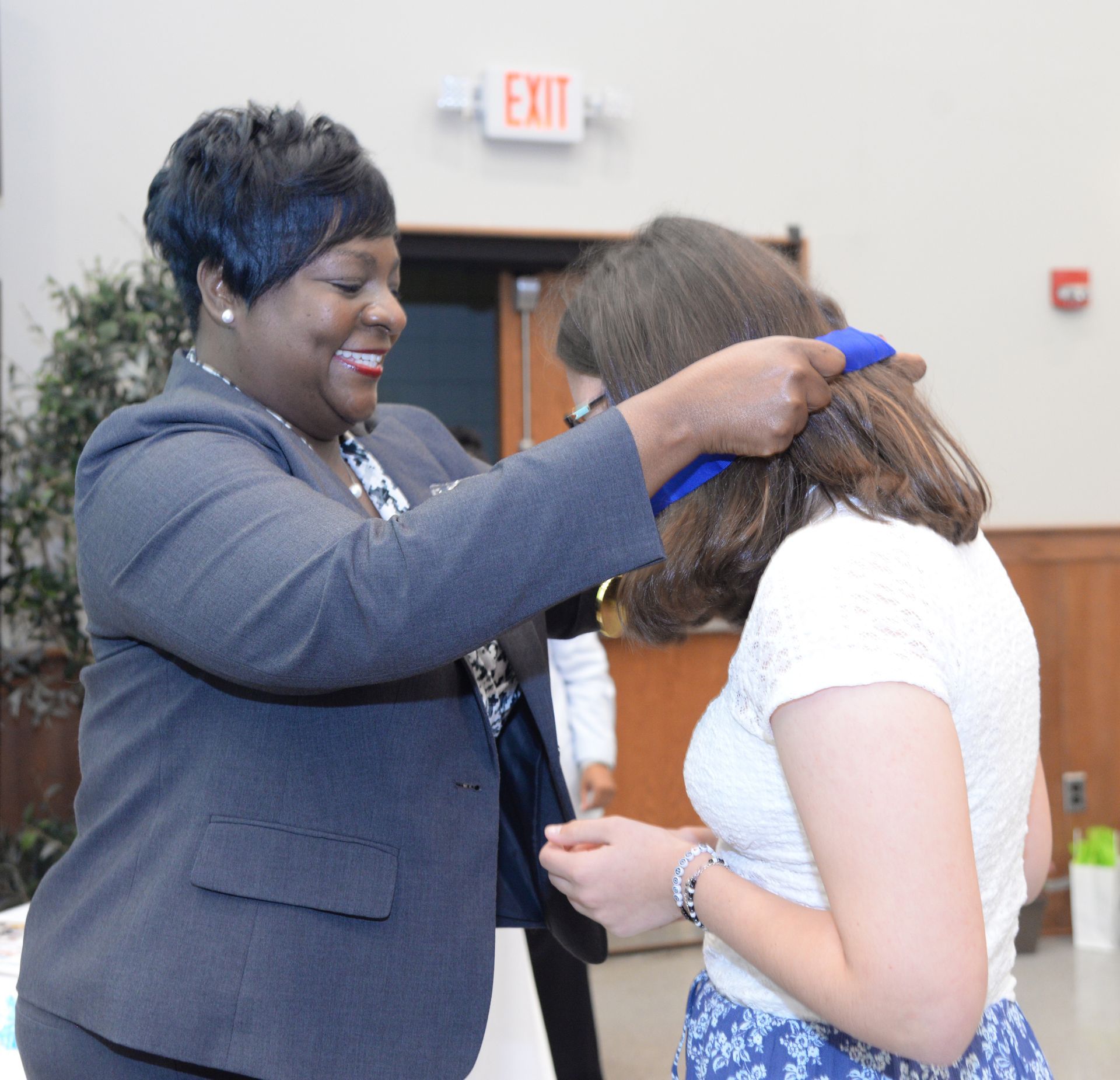 Woman in suit places a blue ribbon over another woman's head in an event setting.