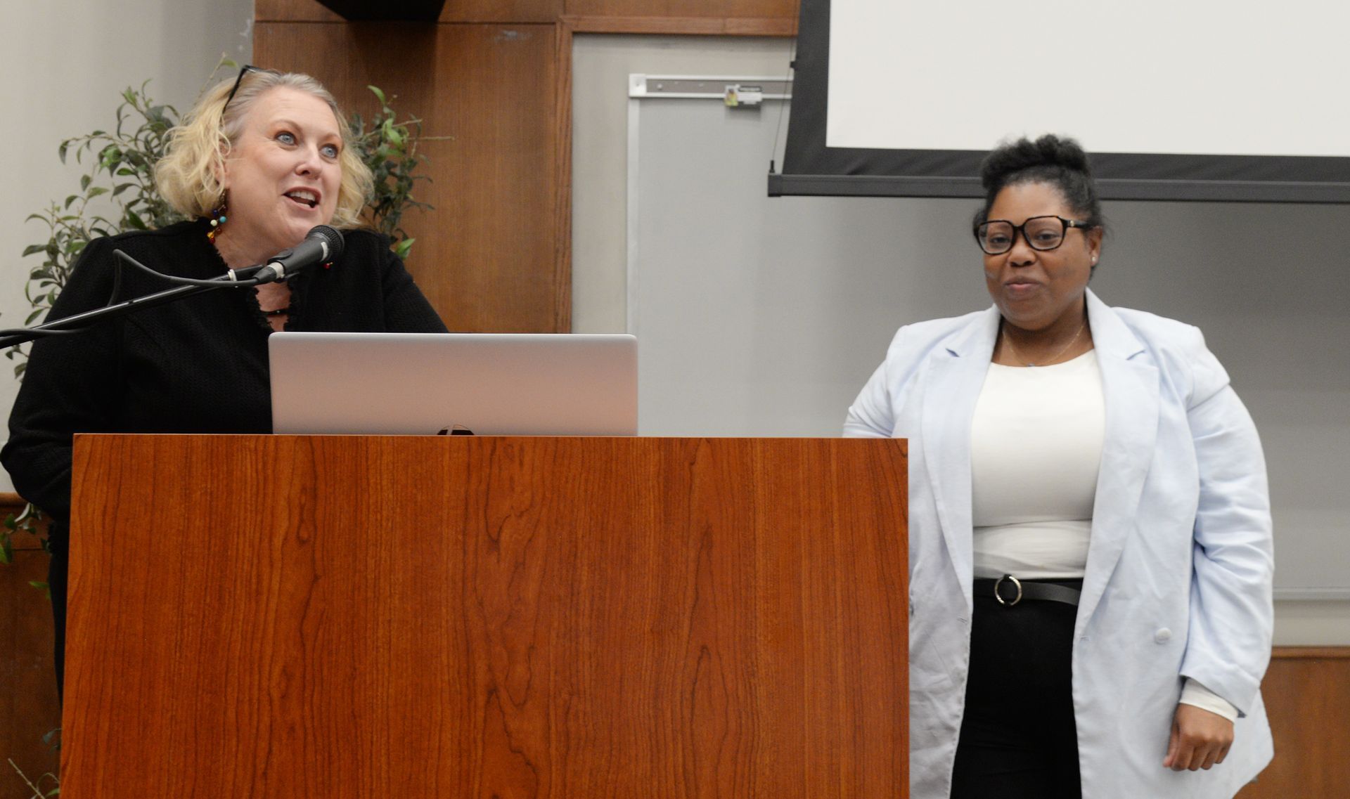 Two women at a wooden podium, one speaking with a microphone, the other standing beside.