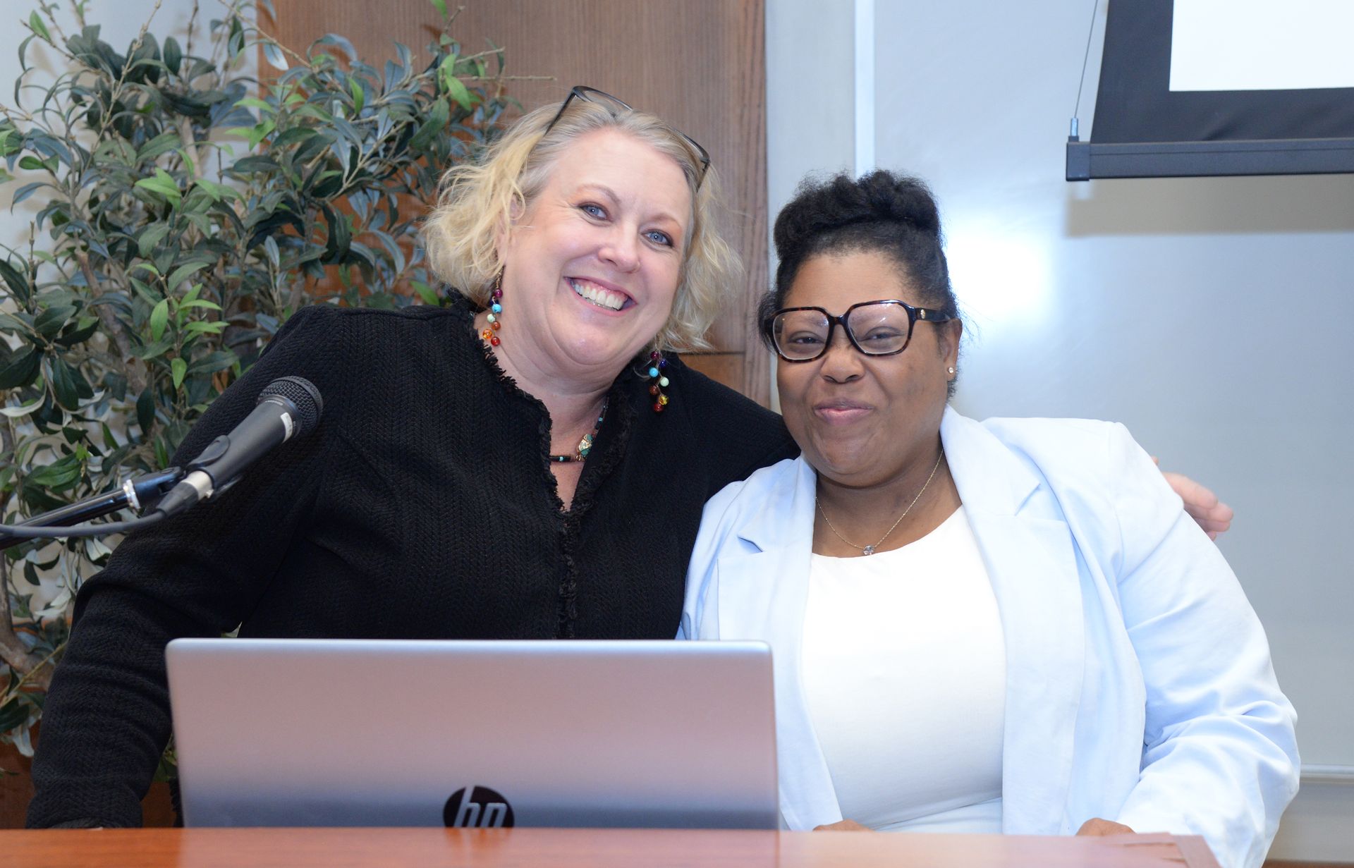 Two smiling people pose by a laptop at a table in a room. One person has blonde hair, the other has dark hair and glasses.