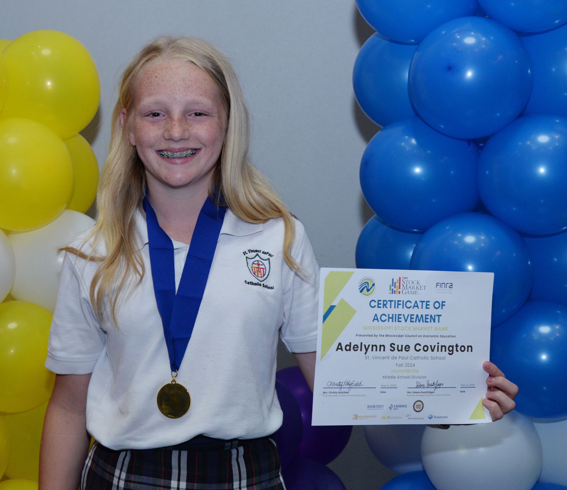 Girl with certificate and medal, smiles at camera. Balloons in background.