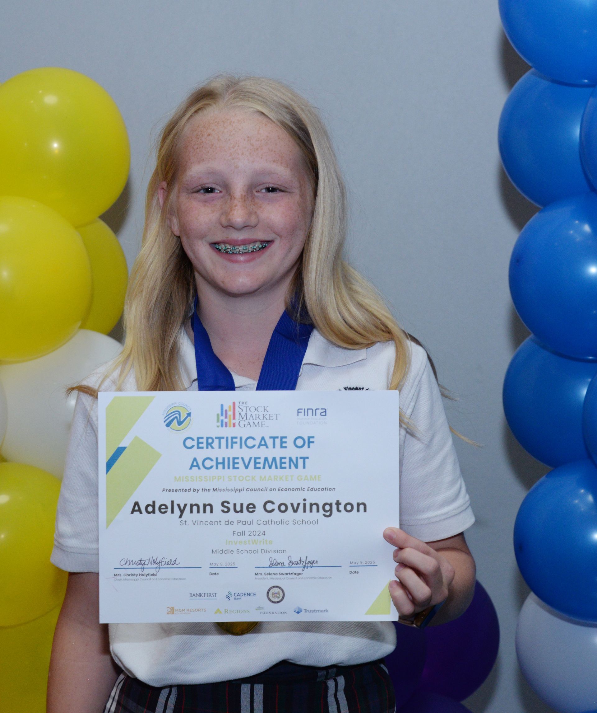 Girl holding a certificate, smiling, with blue and yellow balloons in the background.