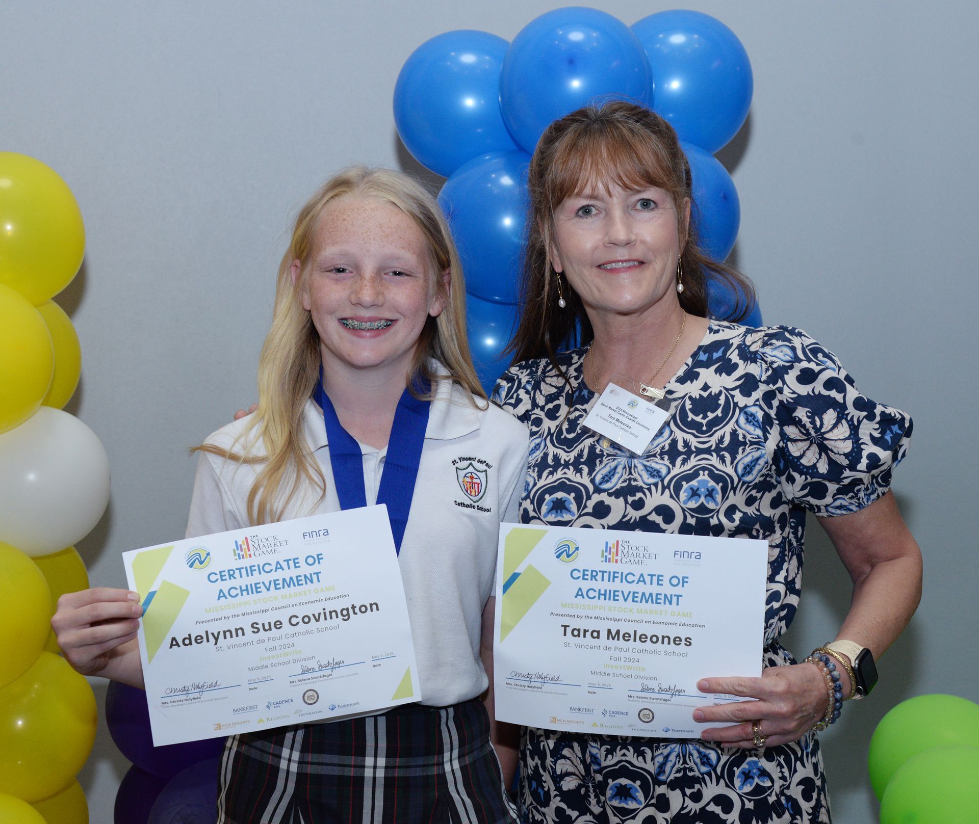 Girl with a medal and woman holding certificates, smiling, next to blue and yellow balloons.