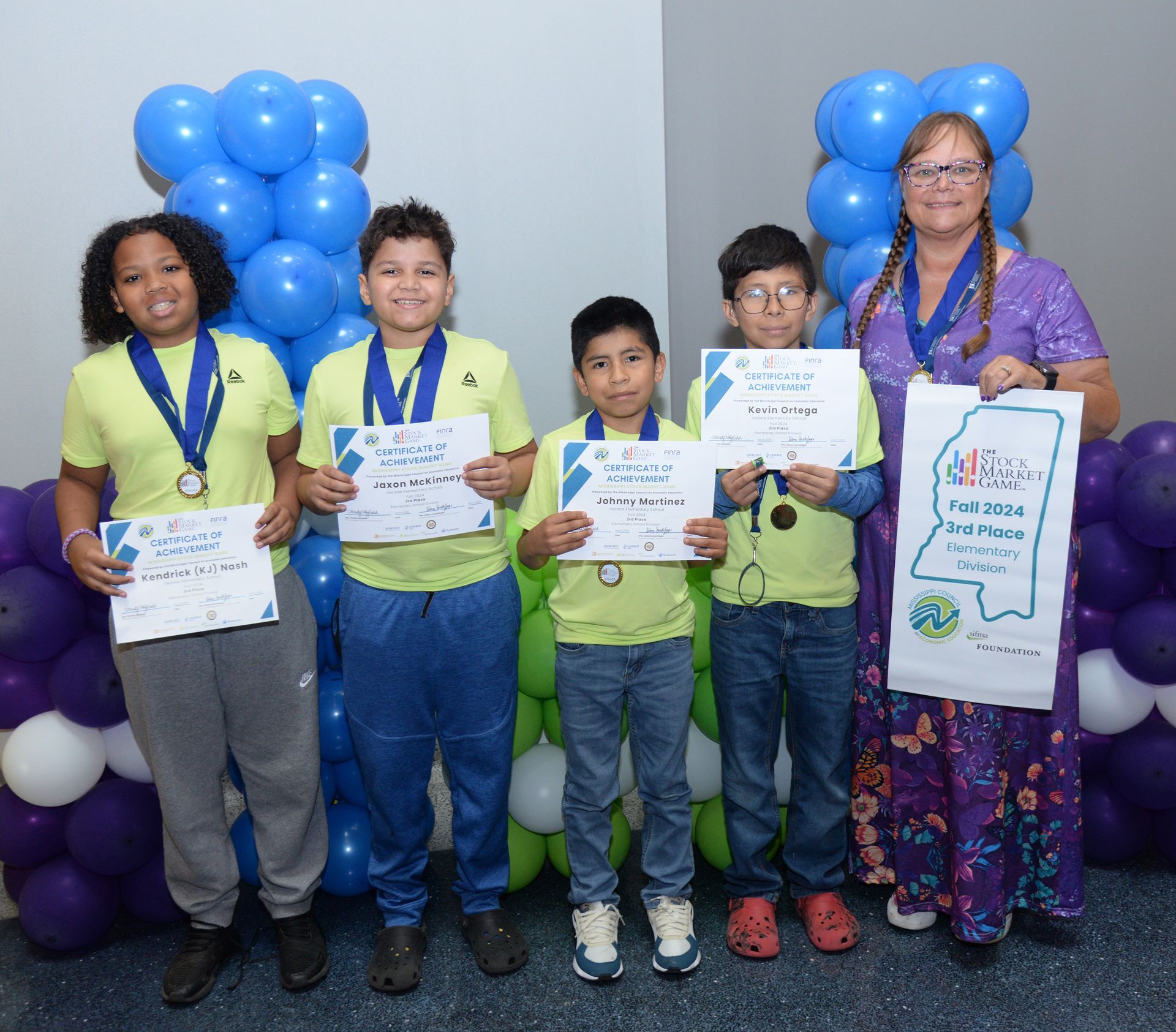 Students hold certificates, medals, and stand with a woman. Balloons in blue, purple, and green.