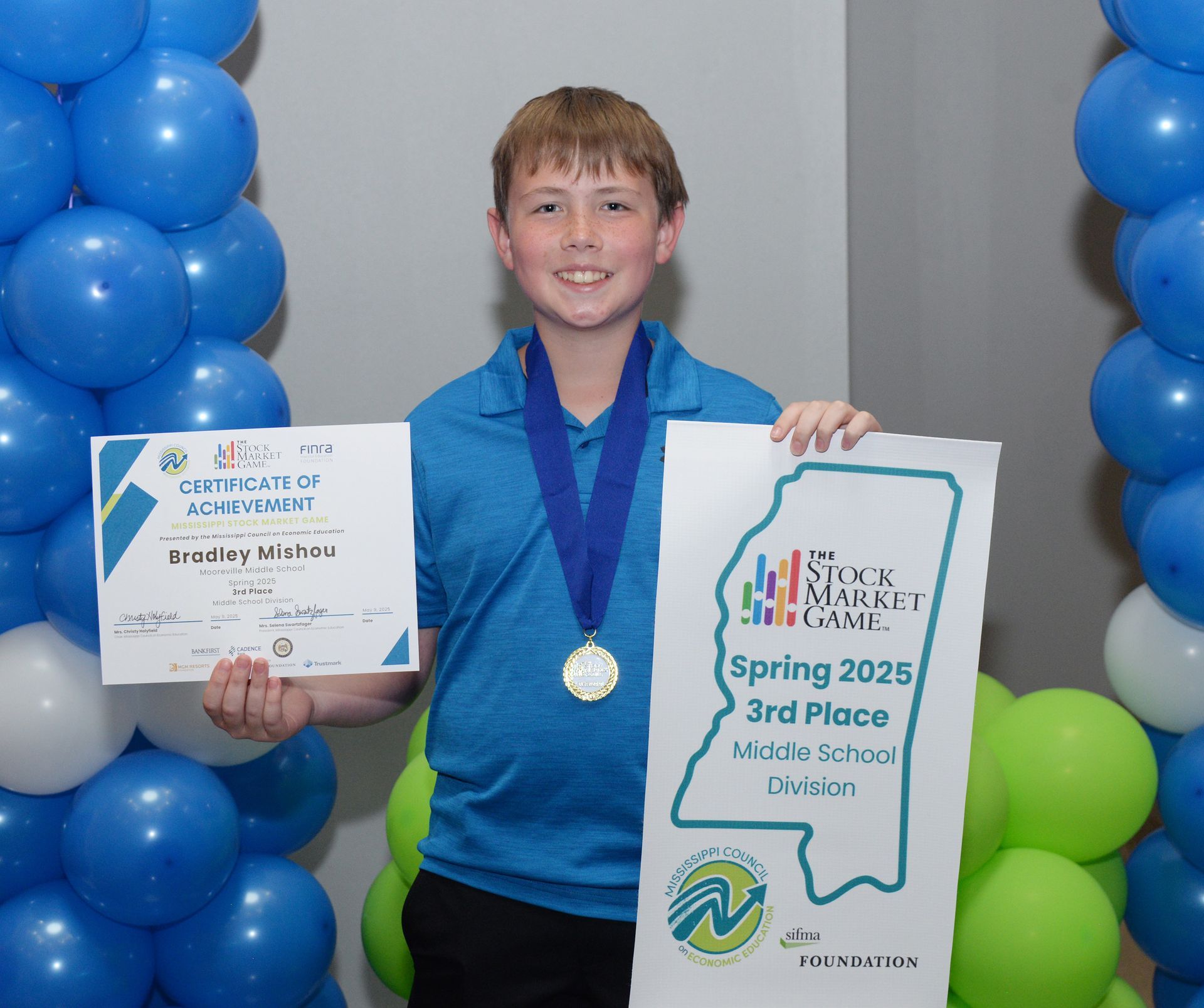 Boy holding certificate and medal, smiling, with blue and green balloons; Stock Market Camp, 3rd place.