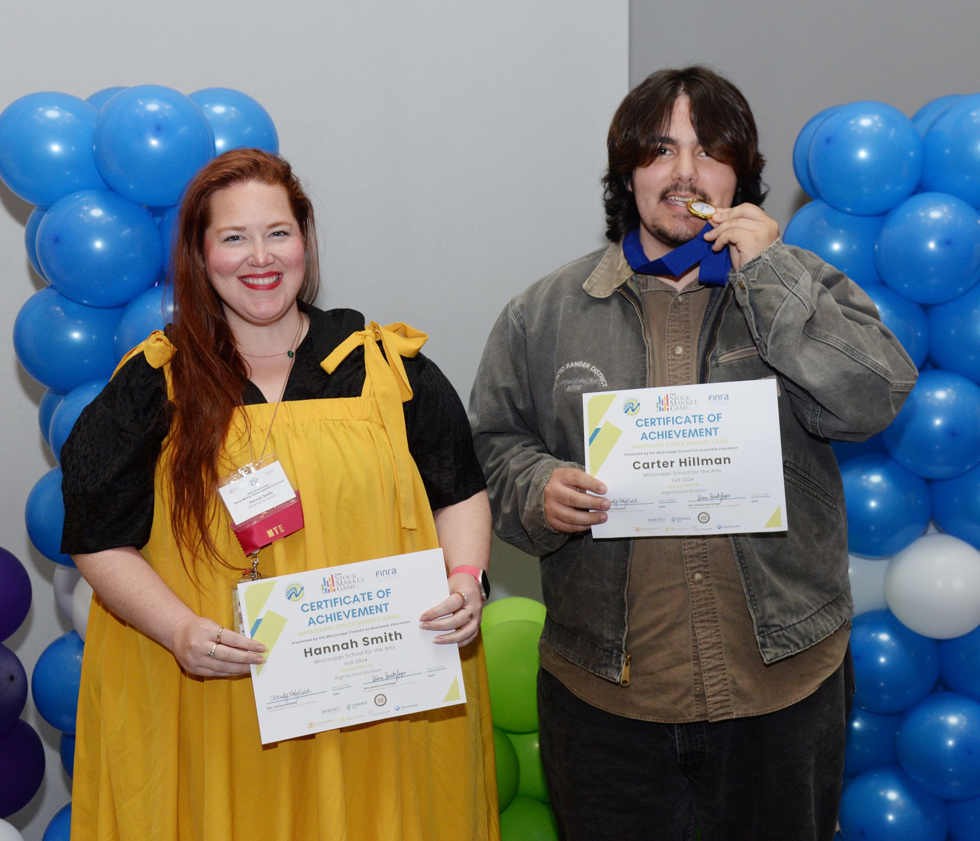 Two people holding certificates, smiling near a blue balloon backdrop. One wears a medal.