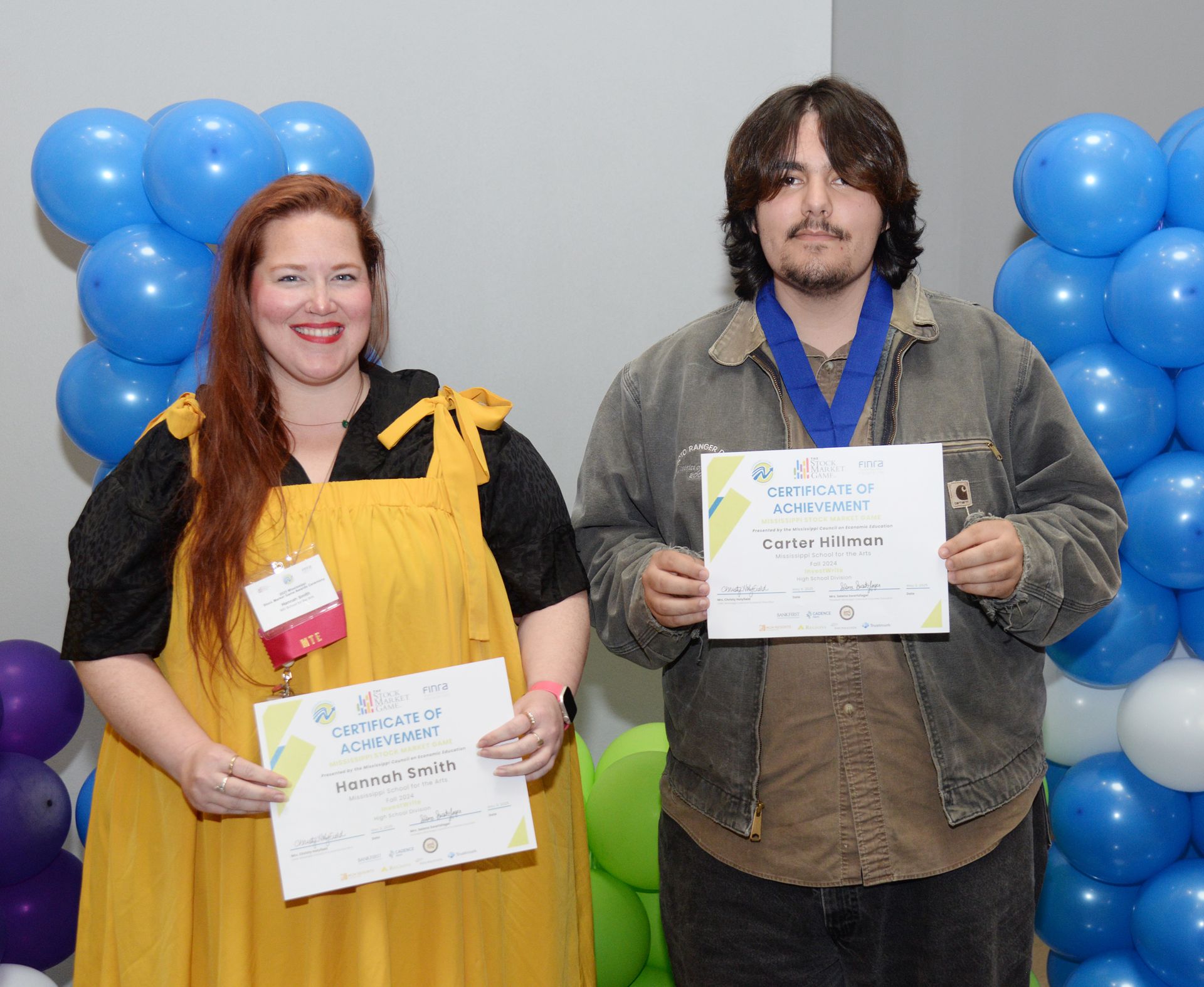 Two people smiling, holding certificates, posing in front of balloons.