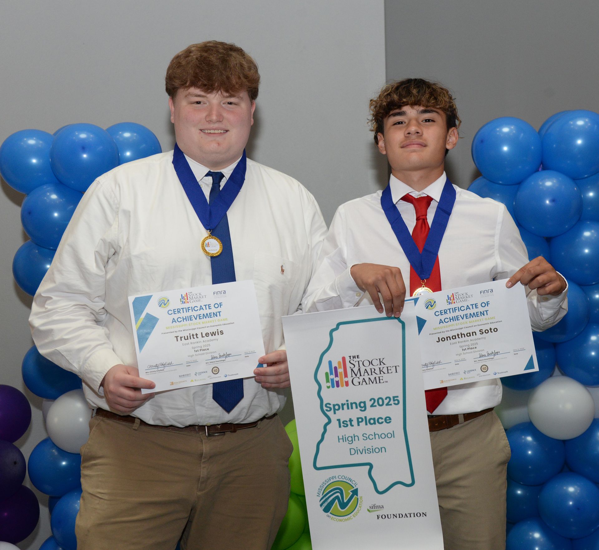 Two young men holding certificates and awards in front of blue balloons.