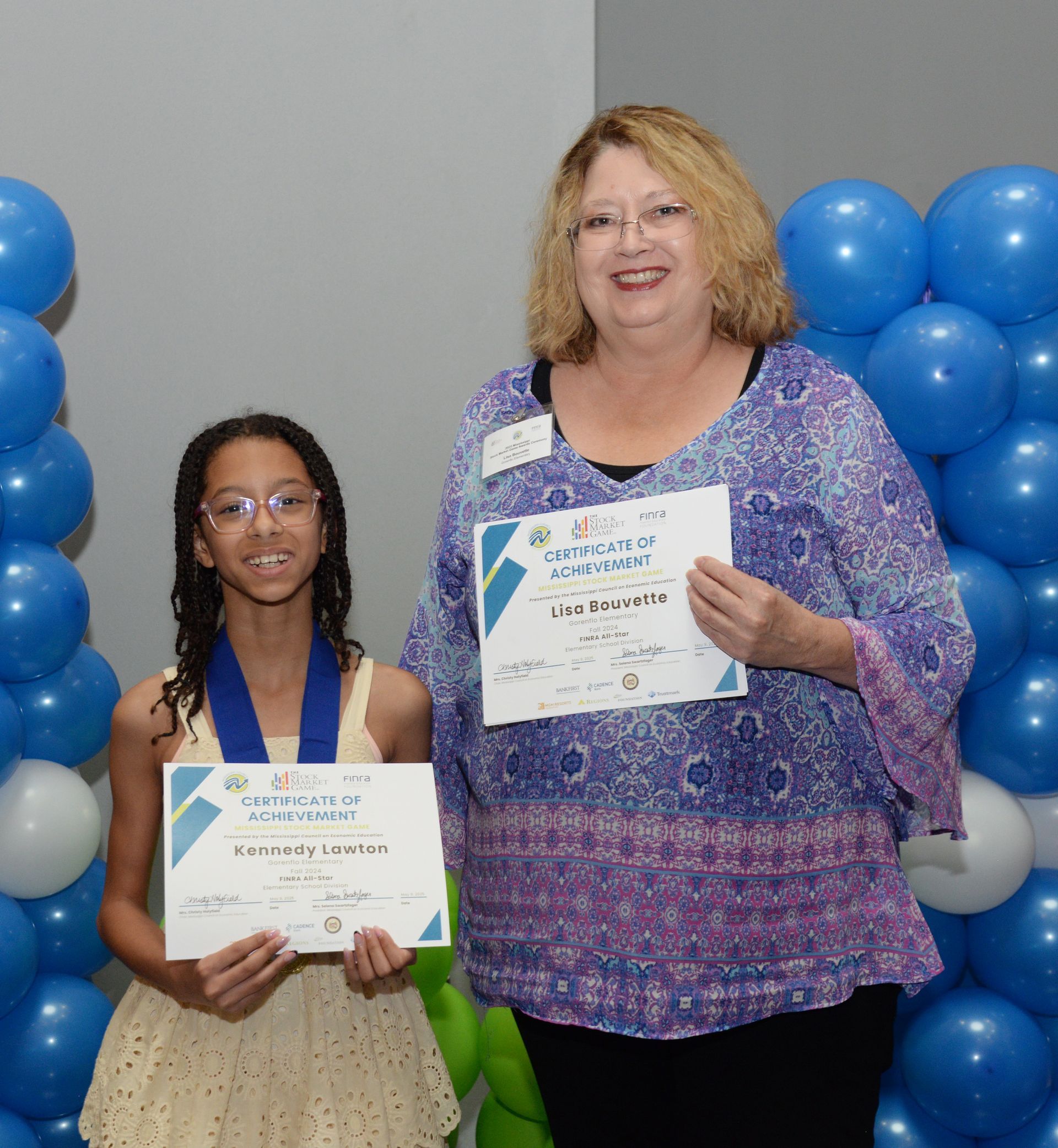 Girl and woman holding certificates, posing in front of balloons, smiling.