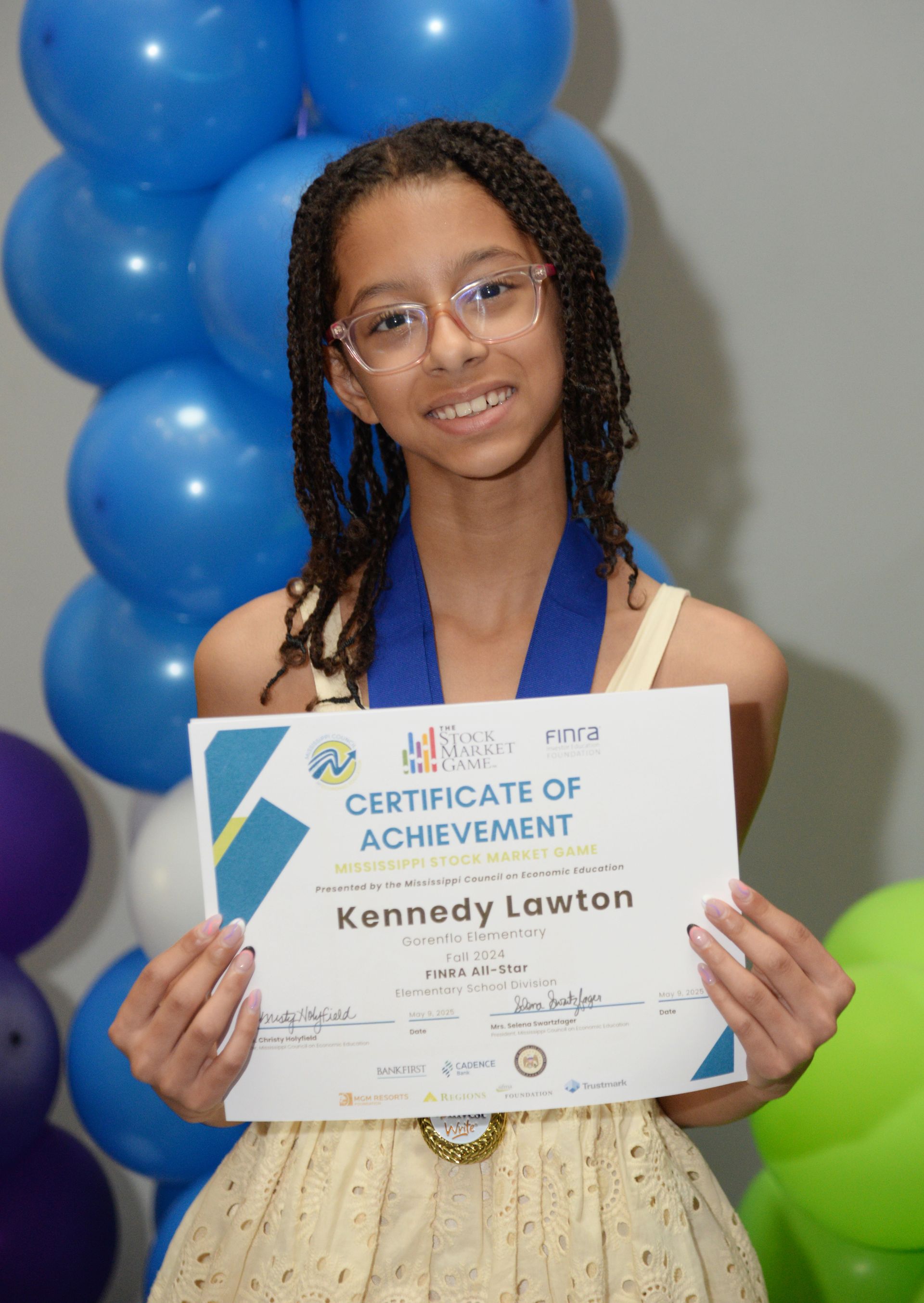Young person holding a certificate, smiling. Wearing glasses and a medal, in front of balloons.