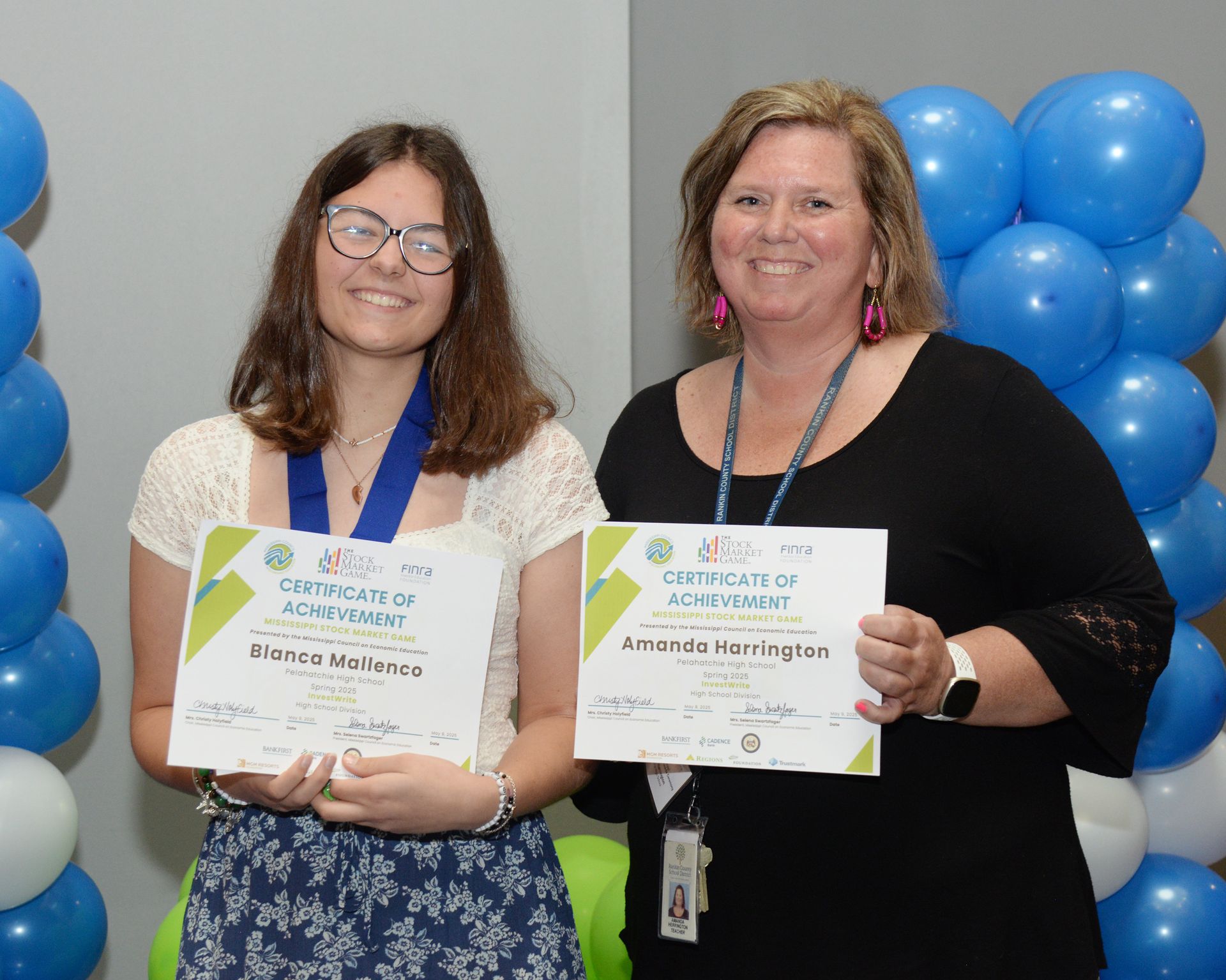 Two women holding certificates, smiling near blue and white balloons.