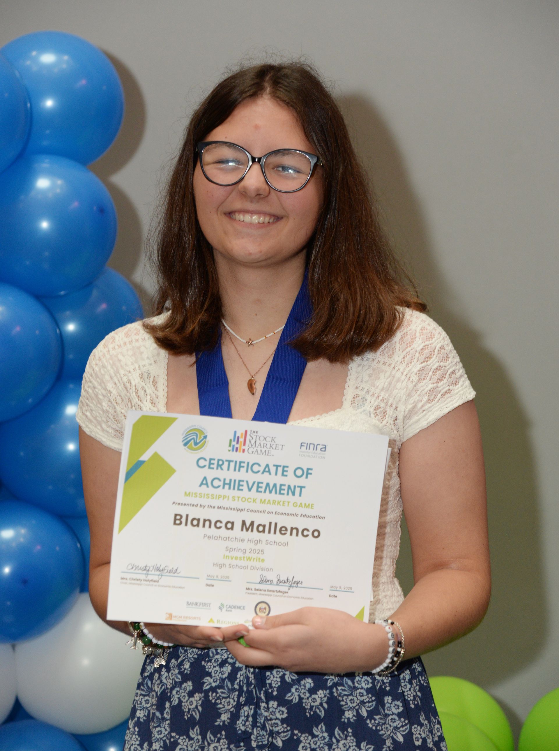 Woman smiles, holding a certificate. Blue and white balloons in the background. She wears glasses and a cream top.