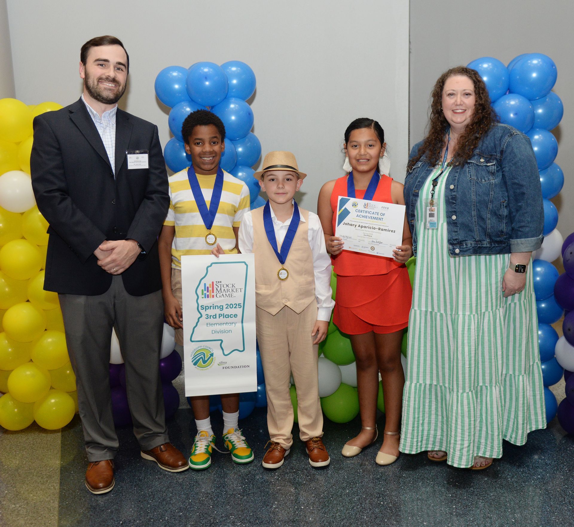 Group posing: adults, children, medals, certificates, balloons, backdrop.