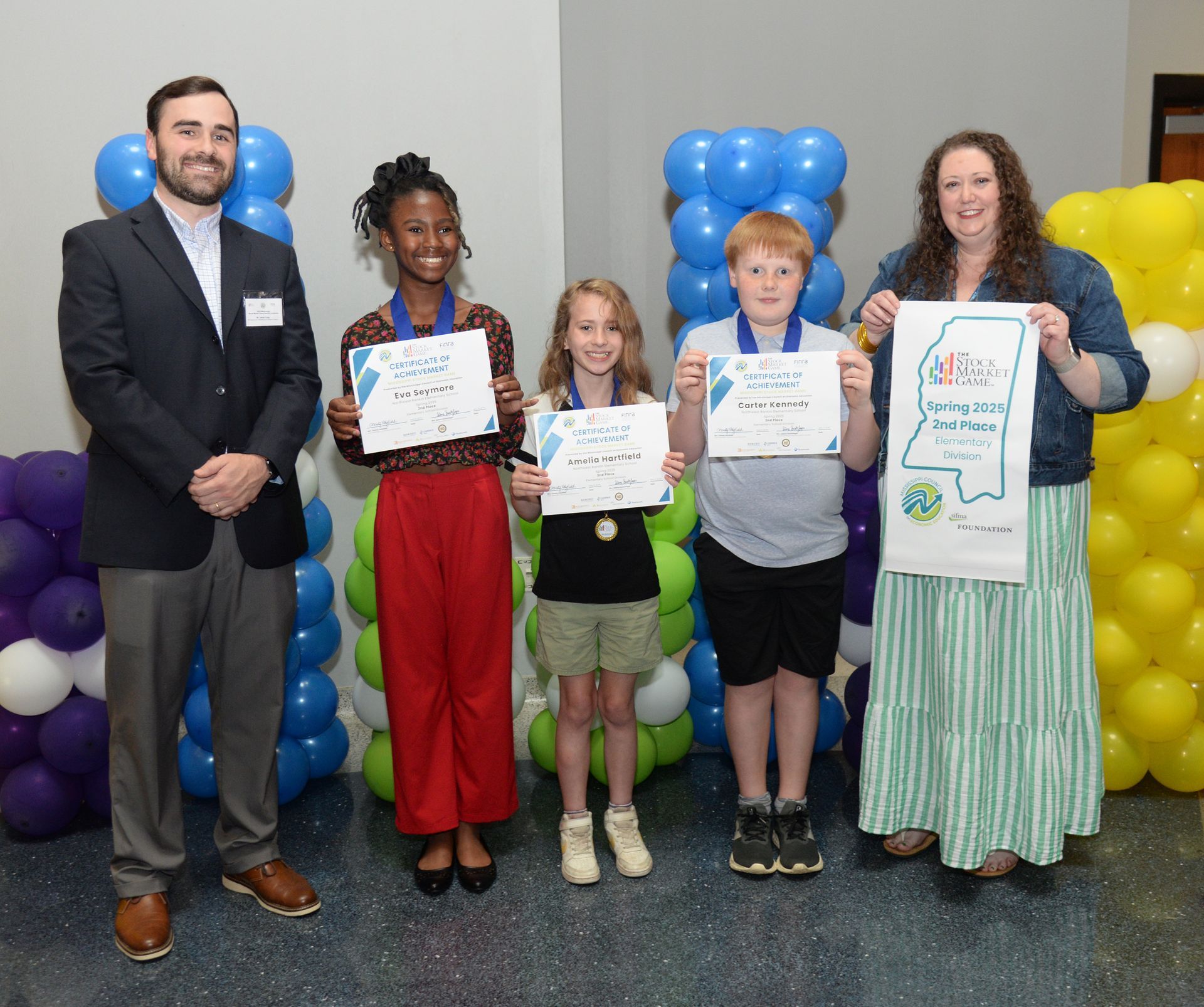 People holding certificates and medals, posing in front of balloon displays.