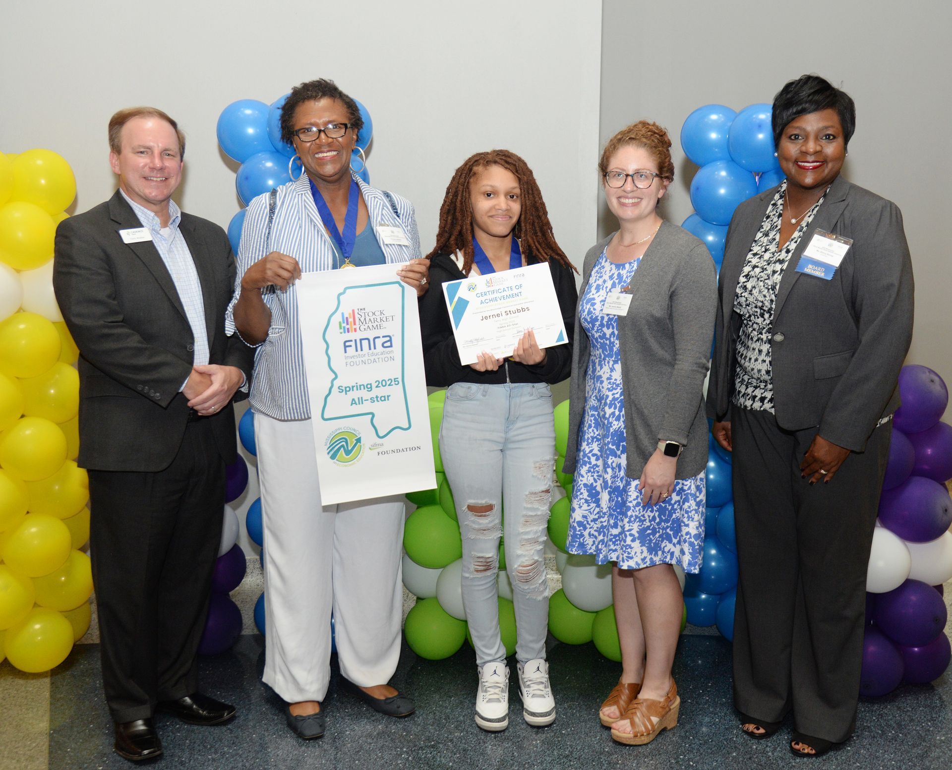 Five people stand near balloons; a teen holds a certificate and bag, others have medals.