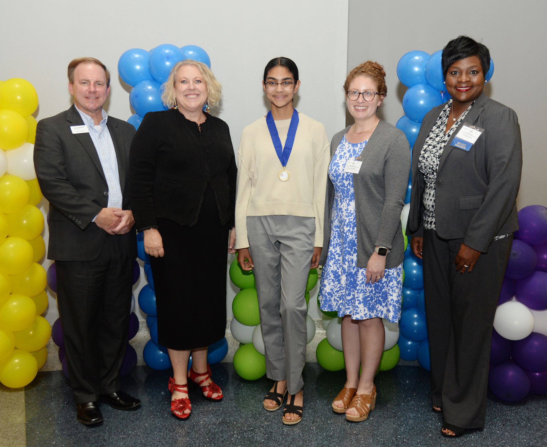 Five people stand together in front of balloon decorations. A person wears a medal; other people wear business attire.