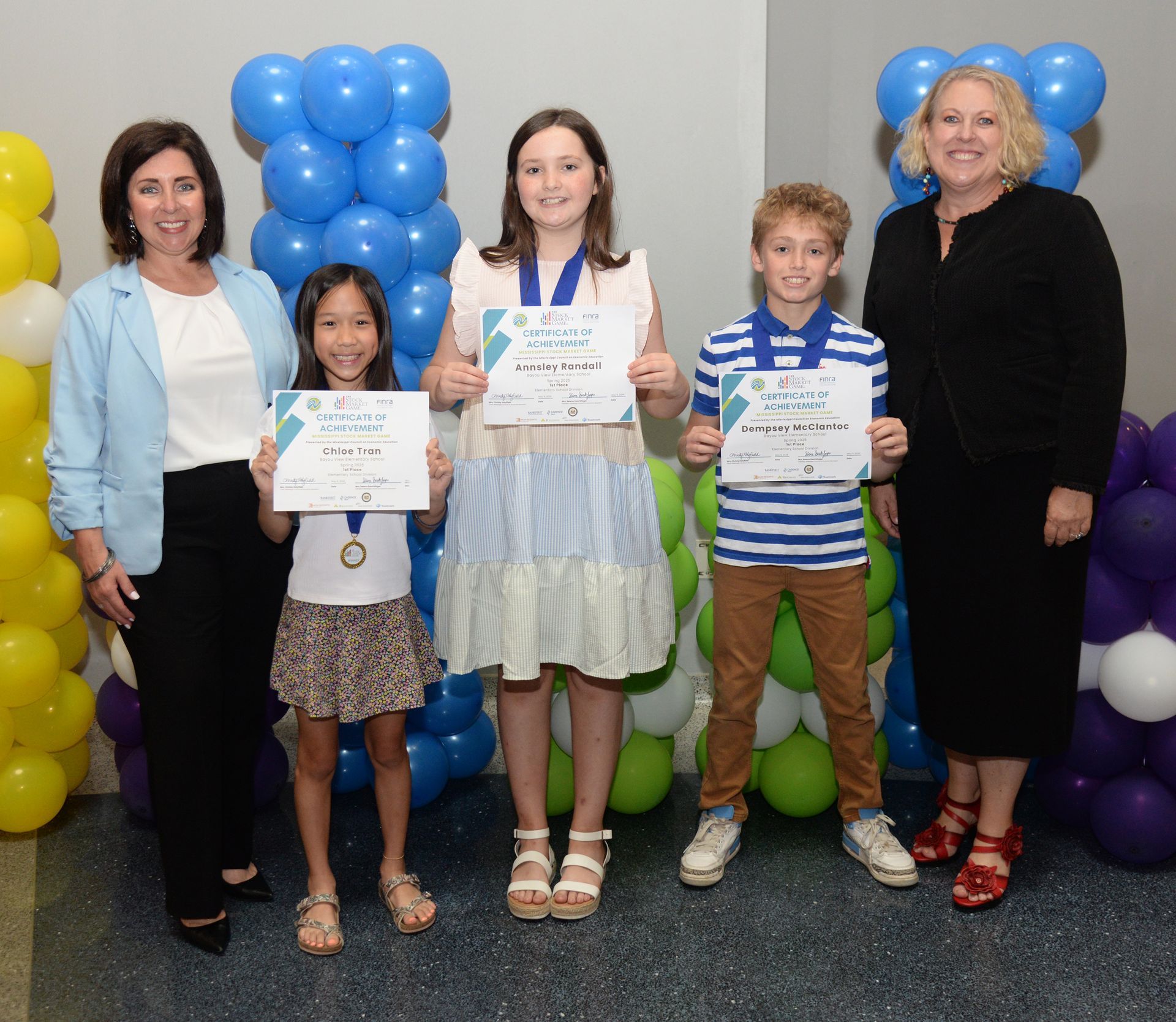 People with certificates and medals, posed with two women, by balloons.