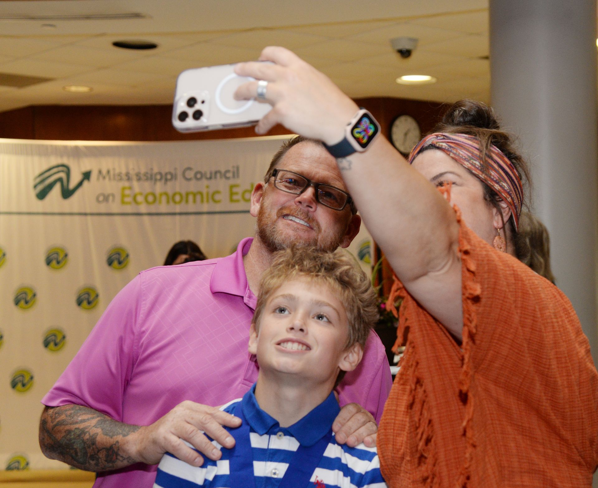 Family takes a selfie together at an event. Man in pink shirt, boy in blue shirt, woman with orange scarf. Background banner.