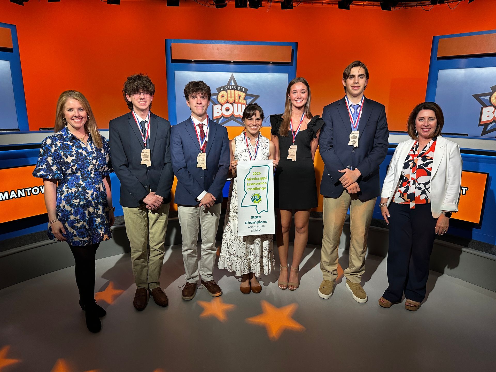 Group of people on a game show stage holding a certificate; orange and blue backdrop; stars on the floor.