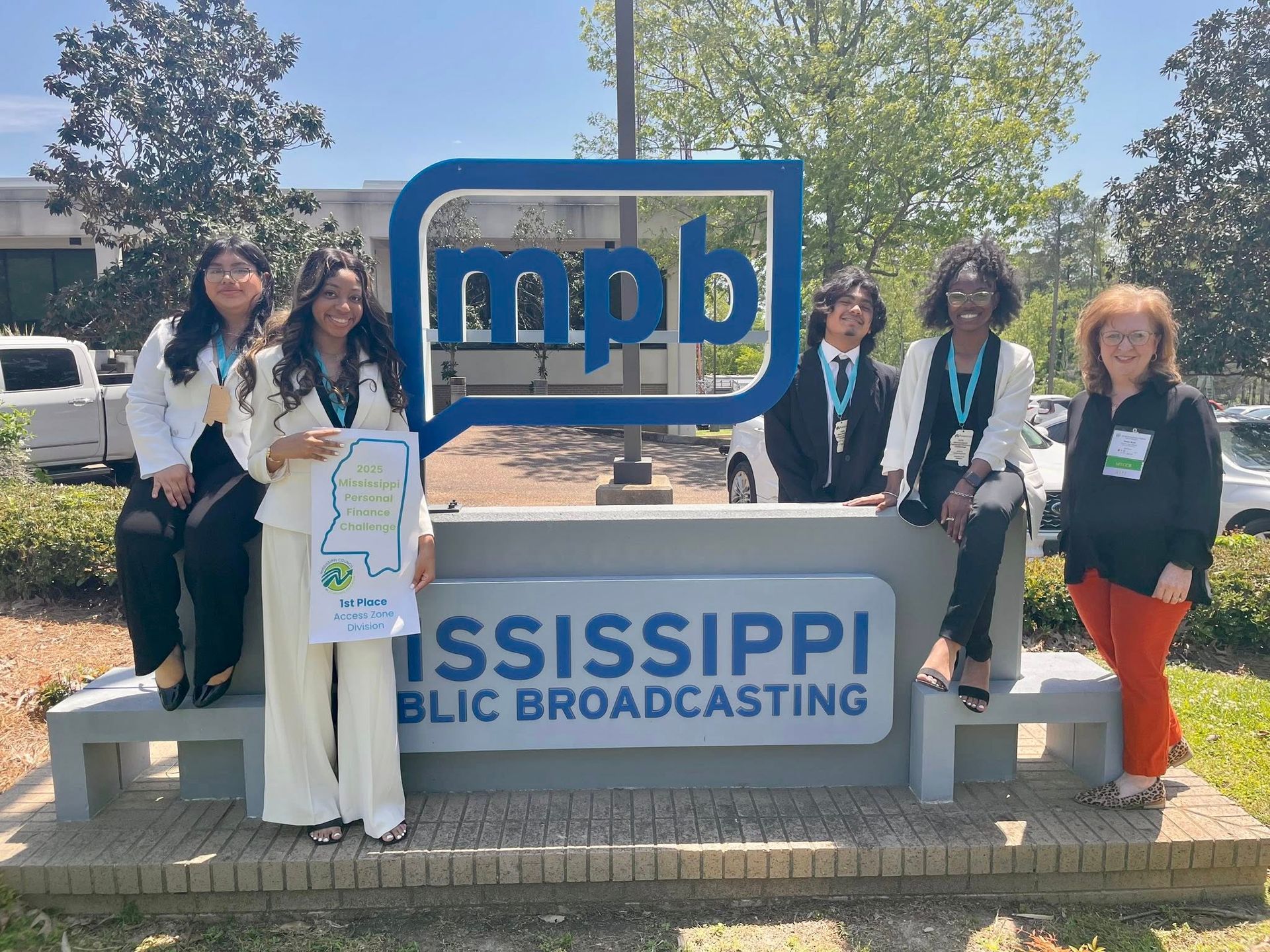 Group poses at the Mississippi Public Broadcasting sign. Holding a sign, they smile in front of the building.