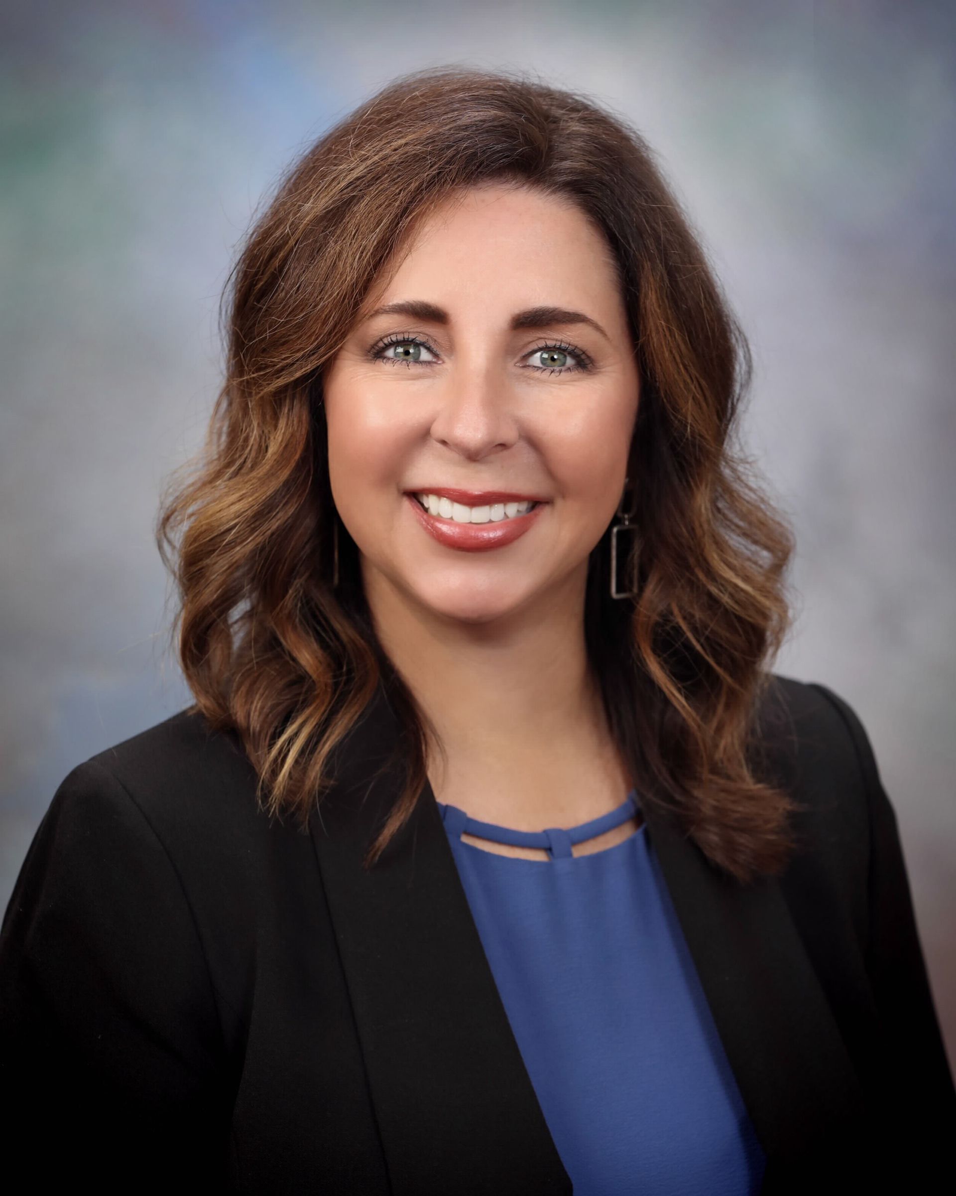 Woman smiling, wearing a blue blouse and black blazer, with shoulder-length wavy brown hair.