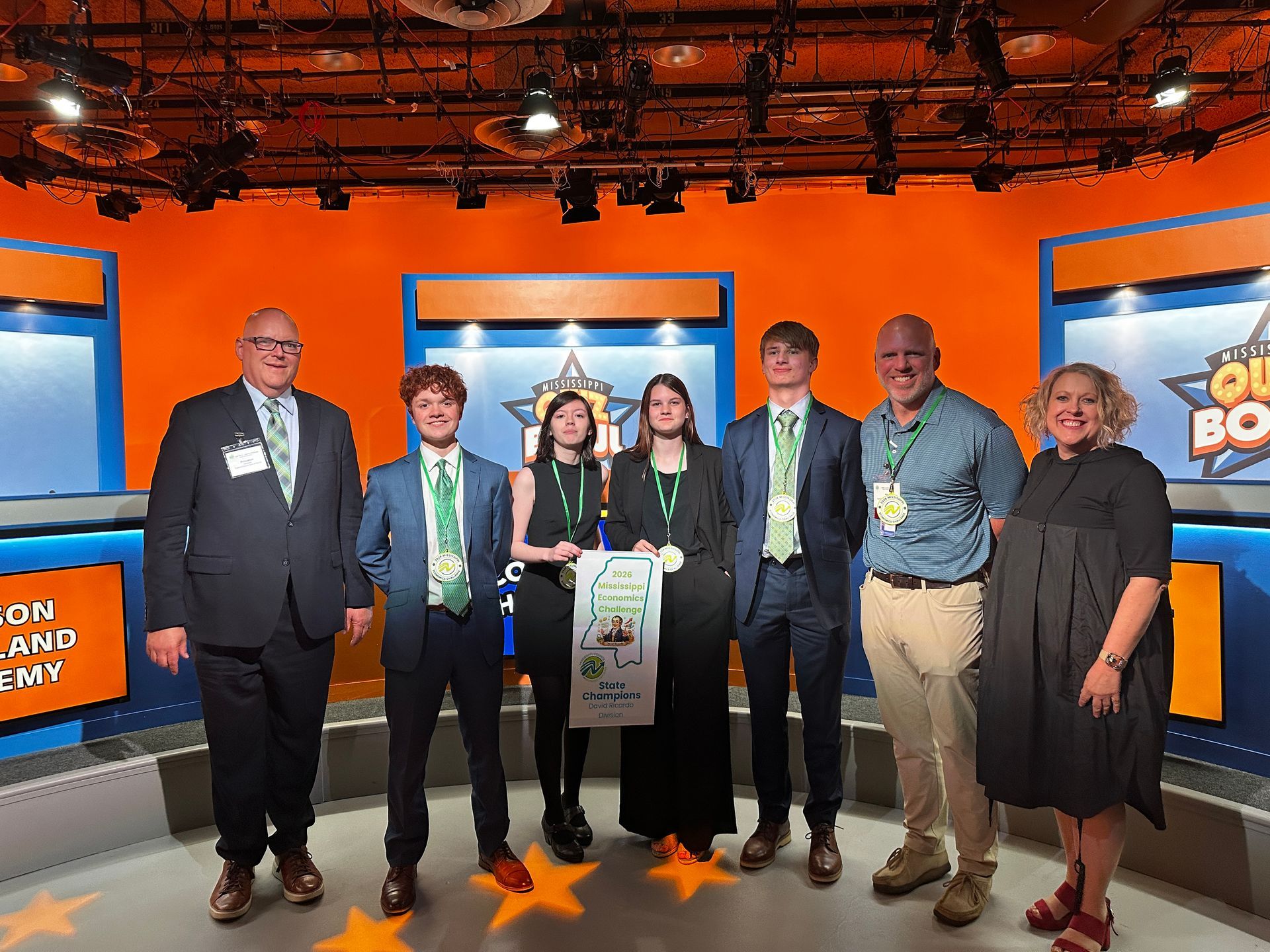 Seven people pose on a bright orange TV studio set with game-show style screens.