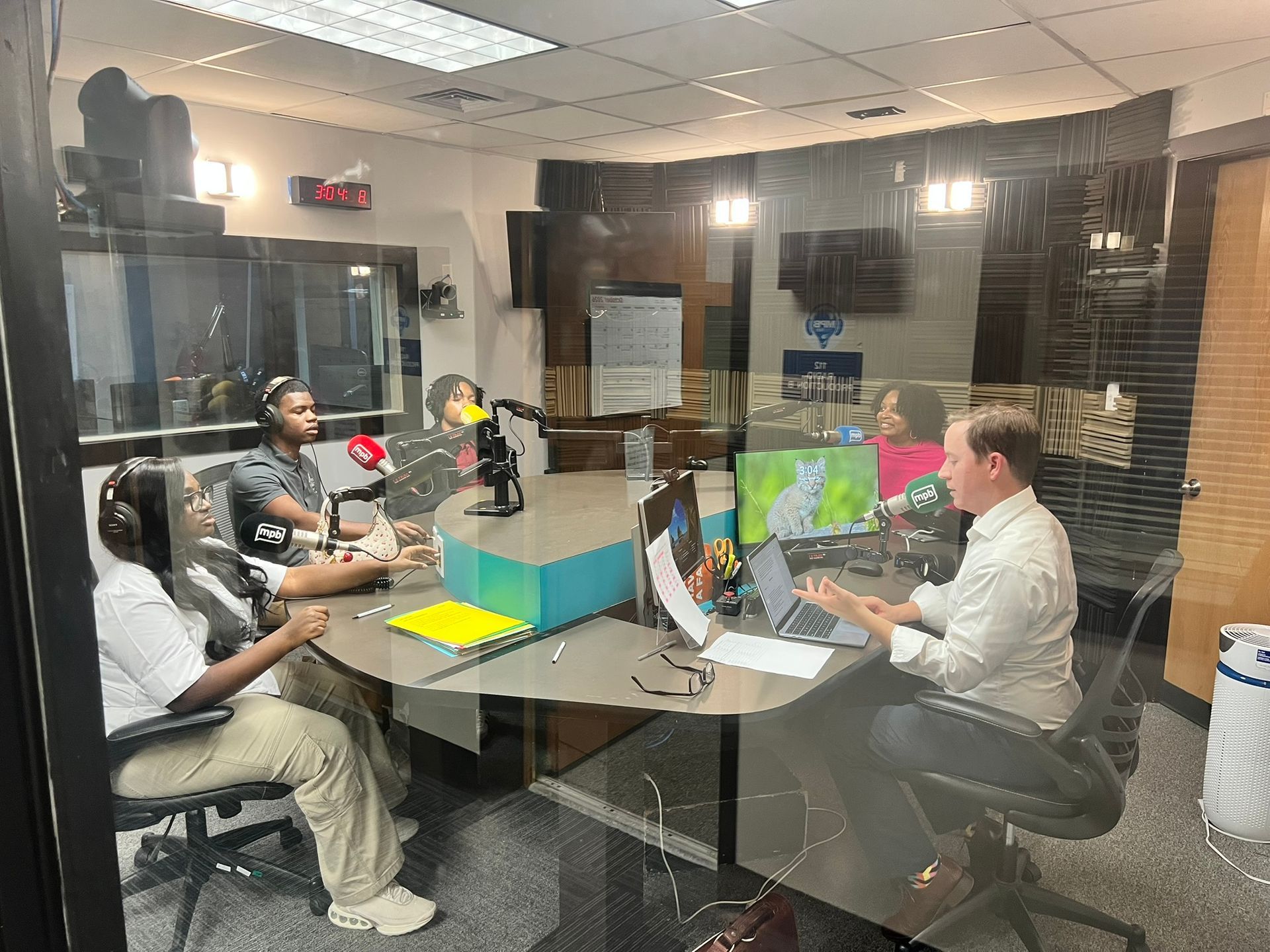 People seated around a table in a glass-walled meeting room, discussing with microphones and laptops.