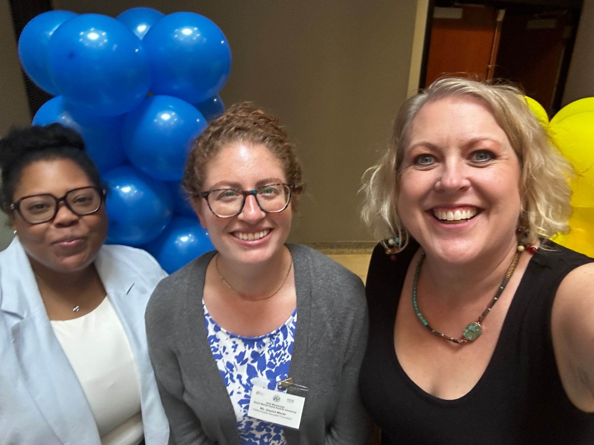 Three smiling people stand near blue balloons in a hallway.