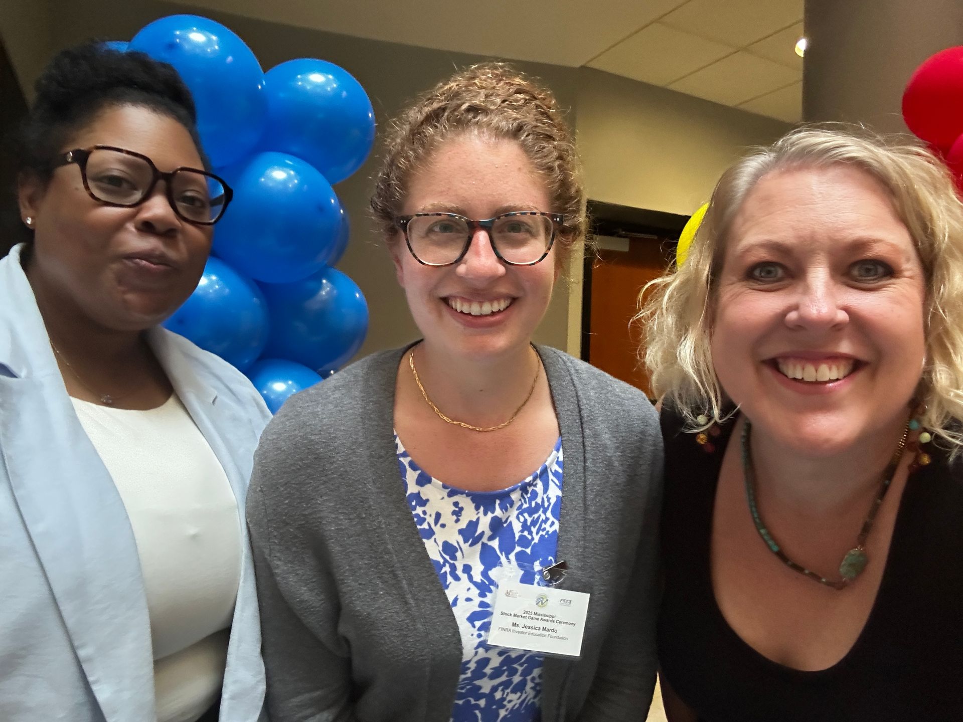 Three smiling people pose in front of blue balloons. Two wear glasses. Indoors, festive setting.