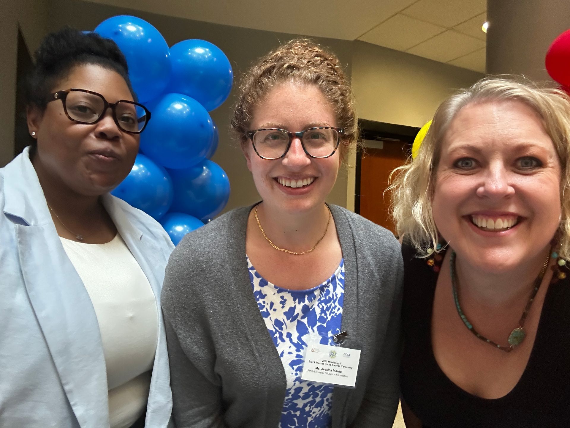 Three people smiling in an indoor setting with blue balloons in the background.