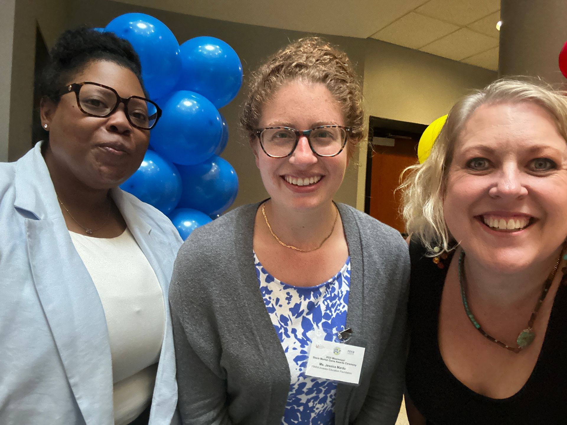 Three people smiling at the camera, blue balloons in the background, a hallway setting.