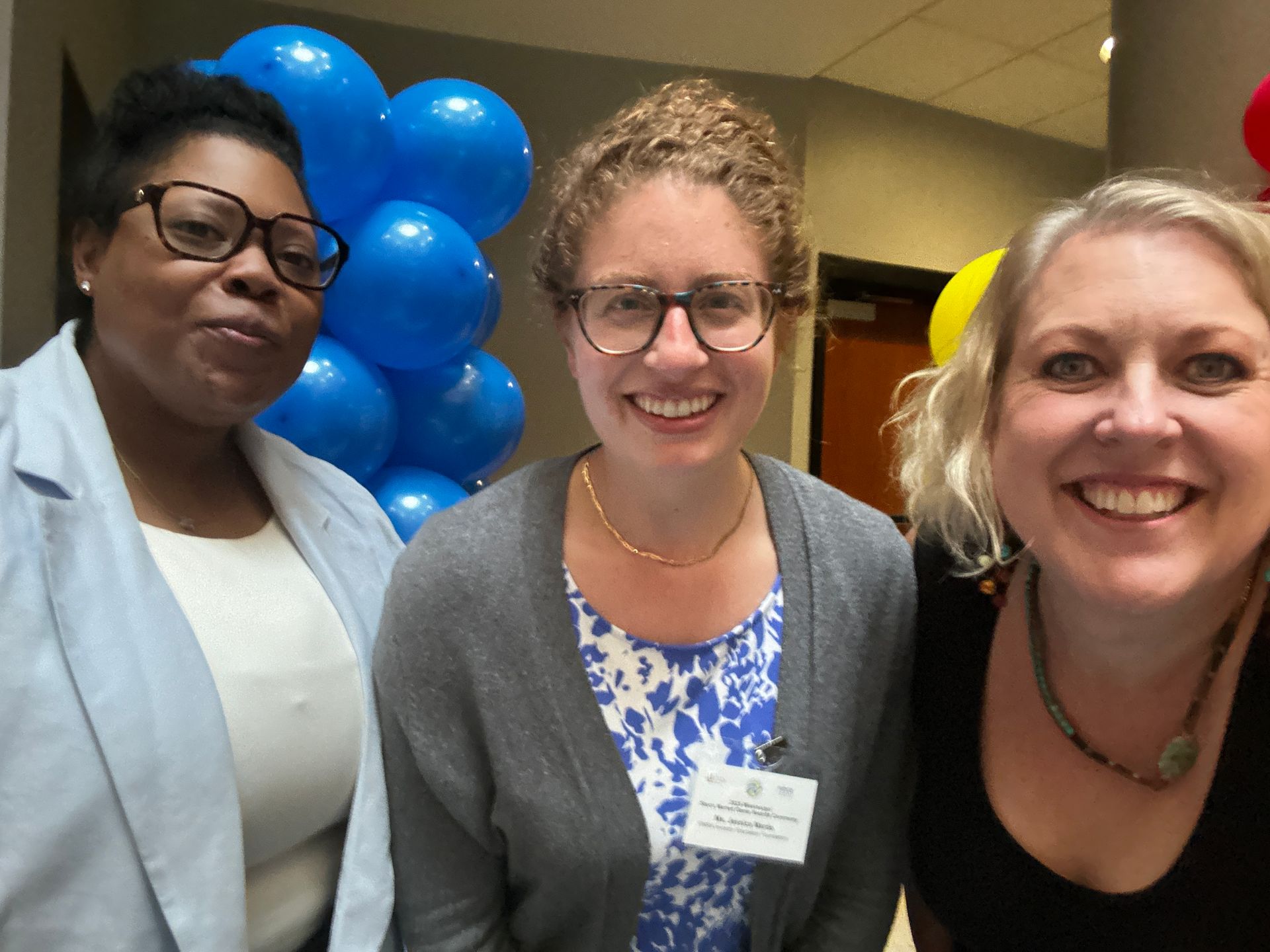 Three smiling people pose in a hallway near blue balloons. Two wear glasses, one a nametag.