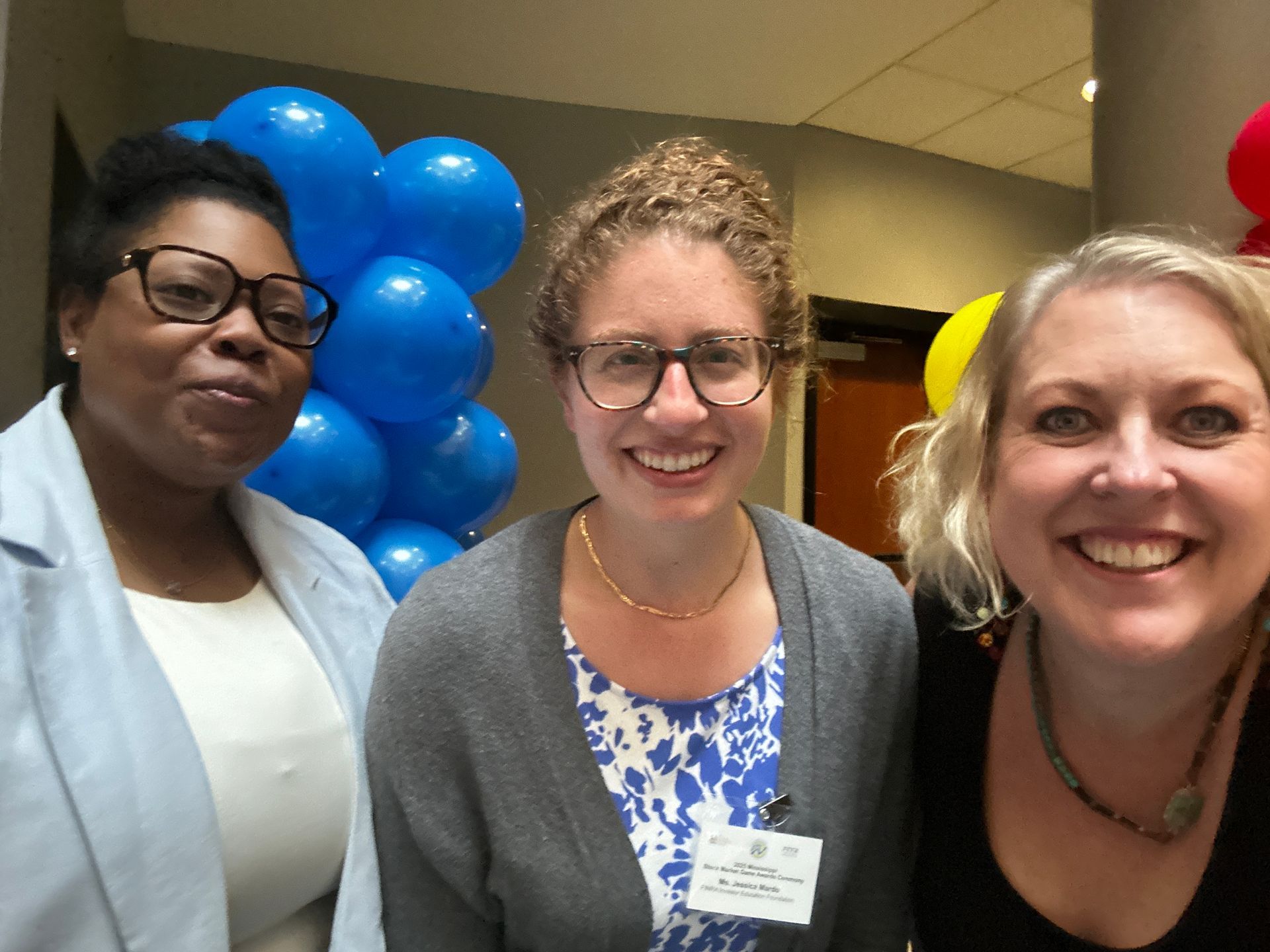 Three smiling people pose in front of blue balloons indoors.